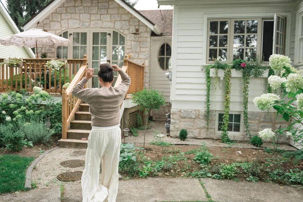 A woman standing in a garden facing away from the camera, adjusting her hair. The background includes a house with a porch, wooden steps, and lush greenery and flowers.