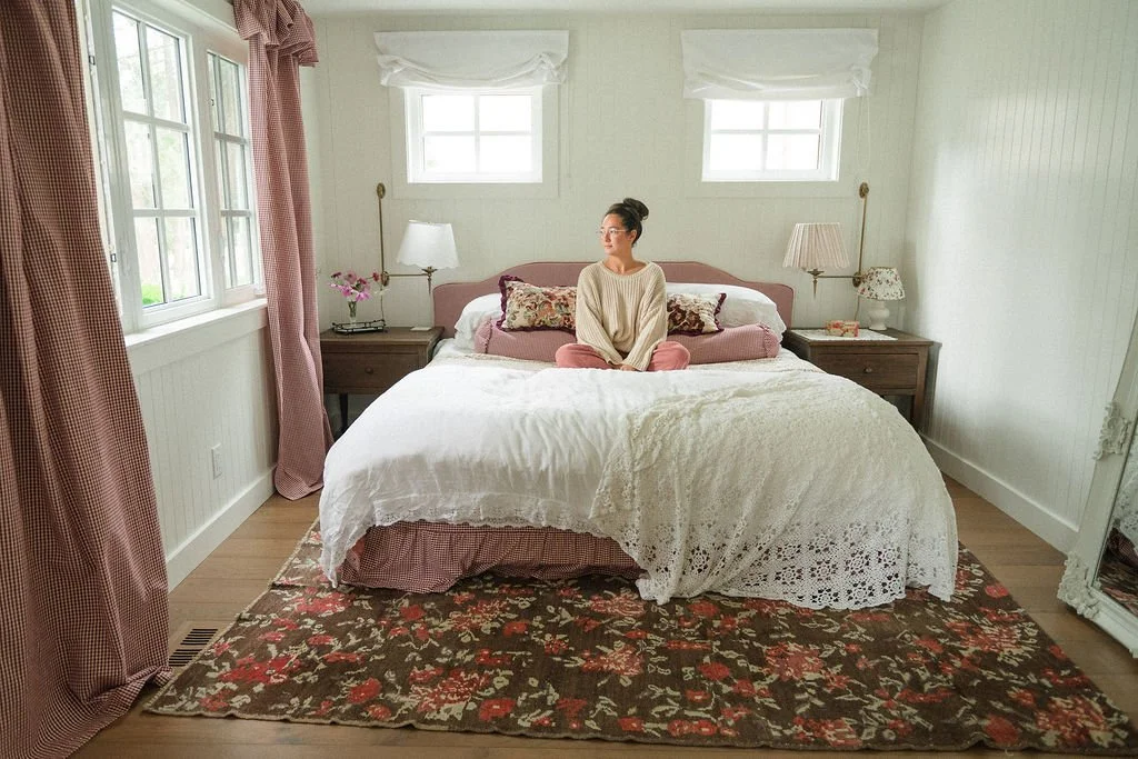 A woman sitting cross-legged on a bed in a bright, cozy room. The bedding is white with decorative pillows. There are two nightstands with lamps on each side of the bed, and pink curtains and valances on the window. An area rug with a floral pattern covers part of the wooden floor.