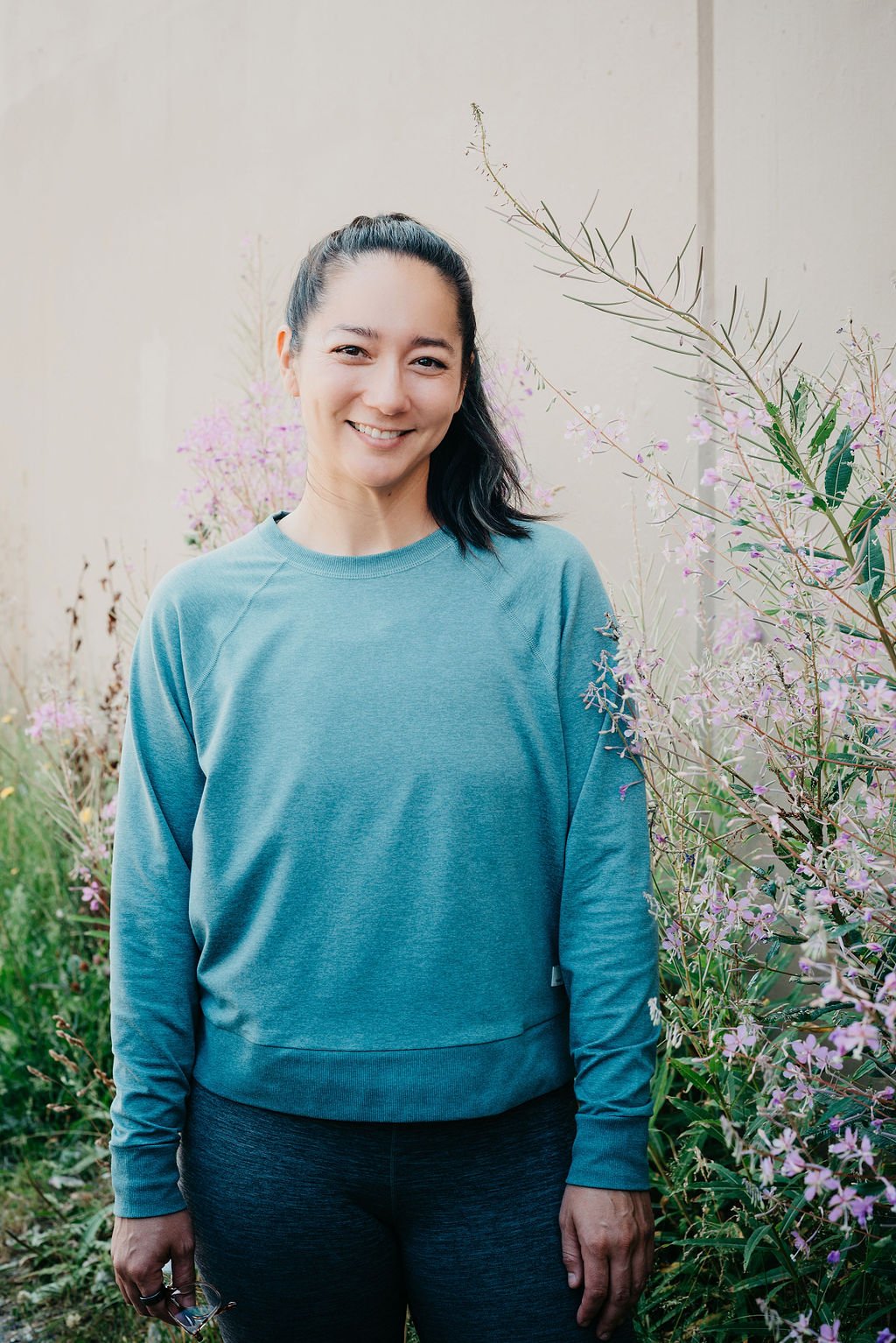 A woman with dark hair in a ponytail, smiling, standing outdoors near pink flowers and greenery, wearing a teal sweatshirt and black pants.