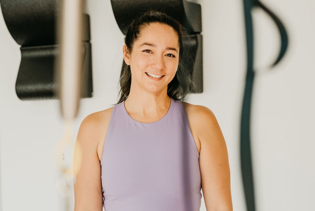A smiling woman in a lavender workout top standing in front of exercise equipment.