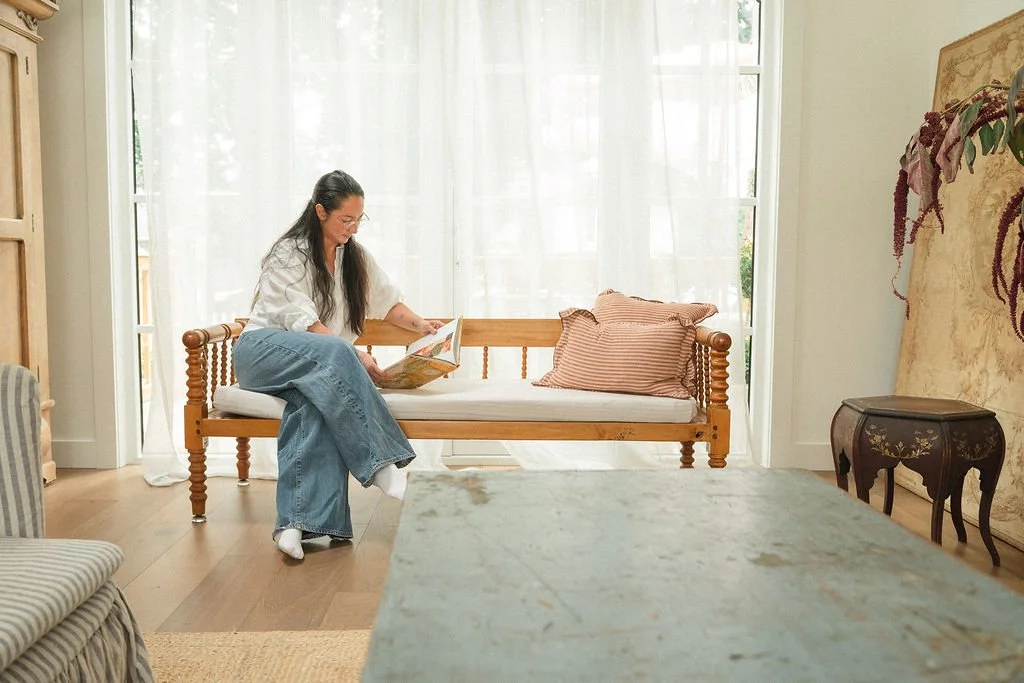 A woman with long dark hair and glasses sitting on a wooden bench, reading a book, in a bright room with white curtains and wooden flooring.