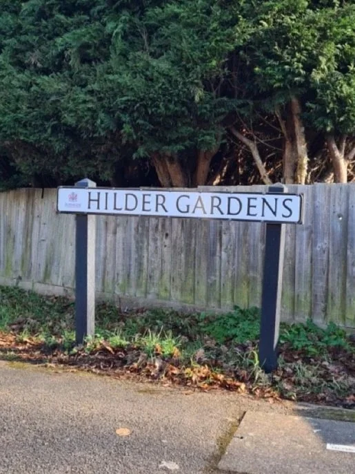 Street sign reading 'HILDER GARDENS' located in front of a wooden fence with trees in the background.