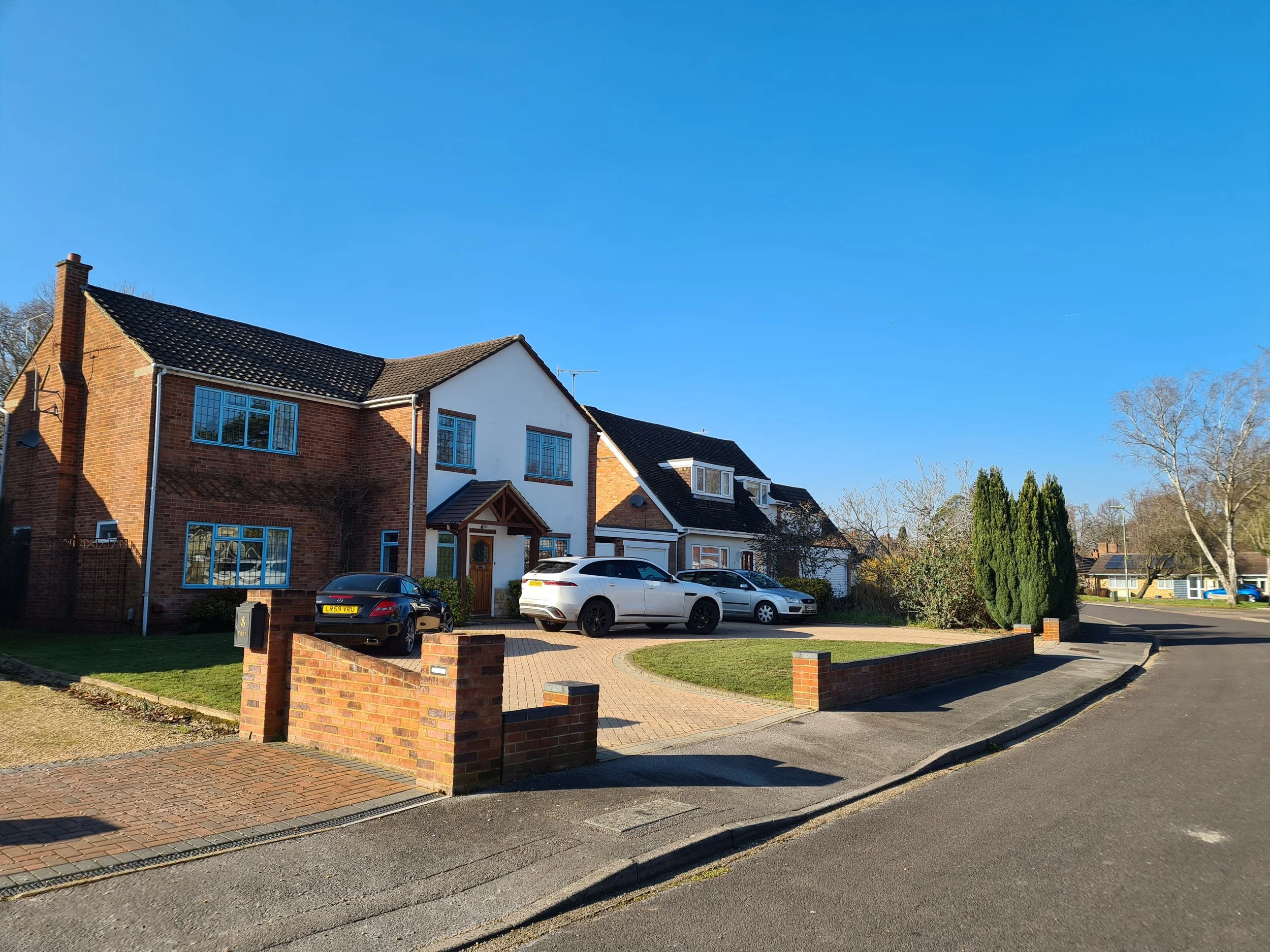 A suburban neighborhood street with houses, cars parked in driveways, a brick wall, and trees under a clear blue sky.