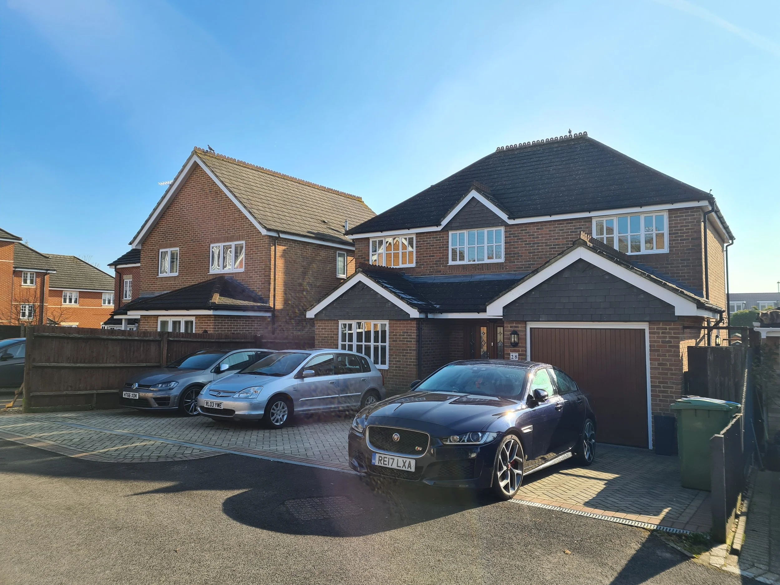 A modern two-story brick house with a dark roof, multiple white-framed windows, and a garage. Four cars are parked in the driveway and front yard, including a black Jaguar, a silver hatchback, and two other vehicles. The sky is clear and blue.
