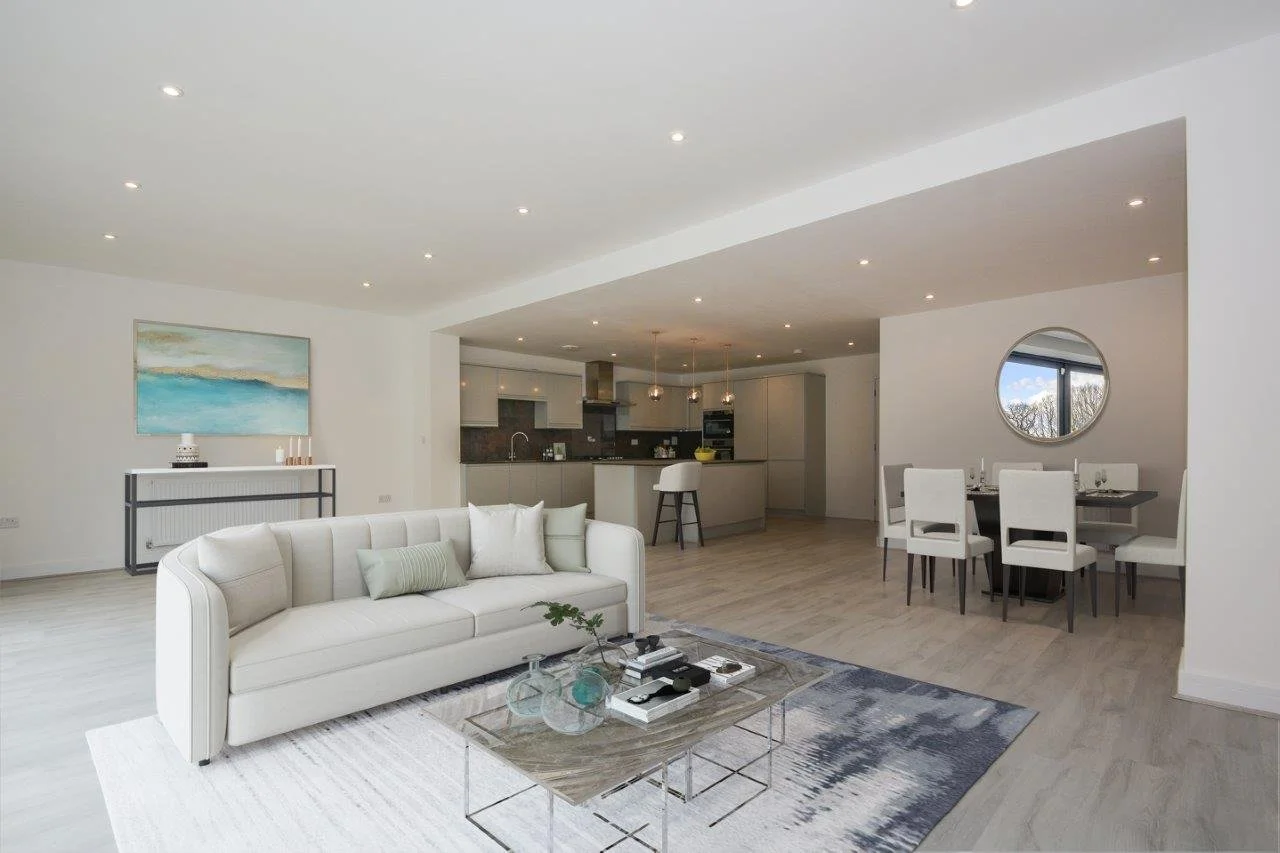Open-plan living room with white sofa, glass coffee table, dining area with table and six chairs, kitchen in the background, round mirror on the wall, and artwork on the wall.