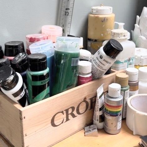 A wooden box filled with various bottles of acrylic paint, jars, and art supplies, with a ruler and additional painting materials visible in the background.