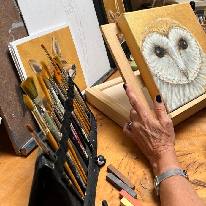 An artist's workspace featuring a hand holding a painting of an owl, brushes in a holder, chalk pastels, and a fox illustration in the background on a wooden table.