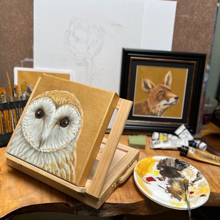 Art studio setup with an owl painting in progress on an easel, a framed fox painting, paint tubes, and a palette with brushes on a table.