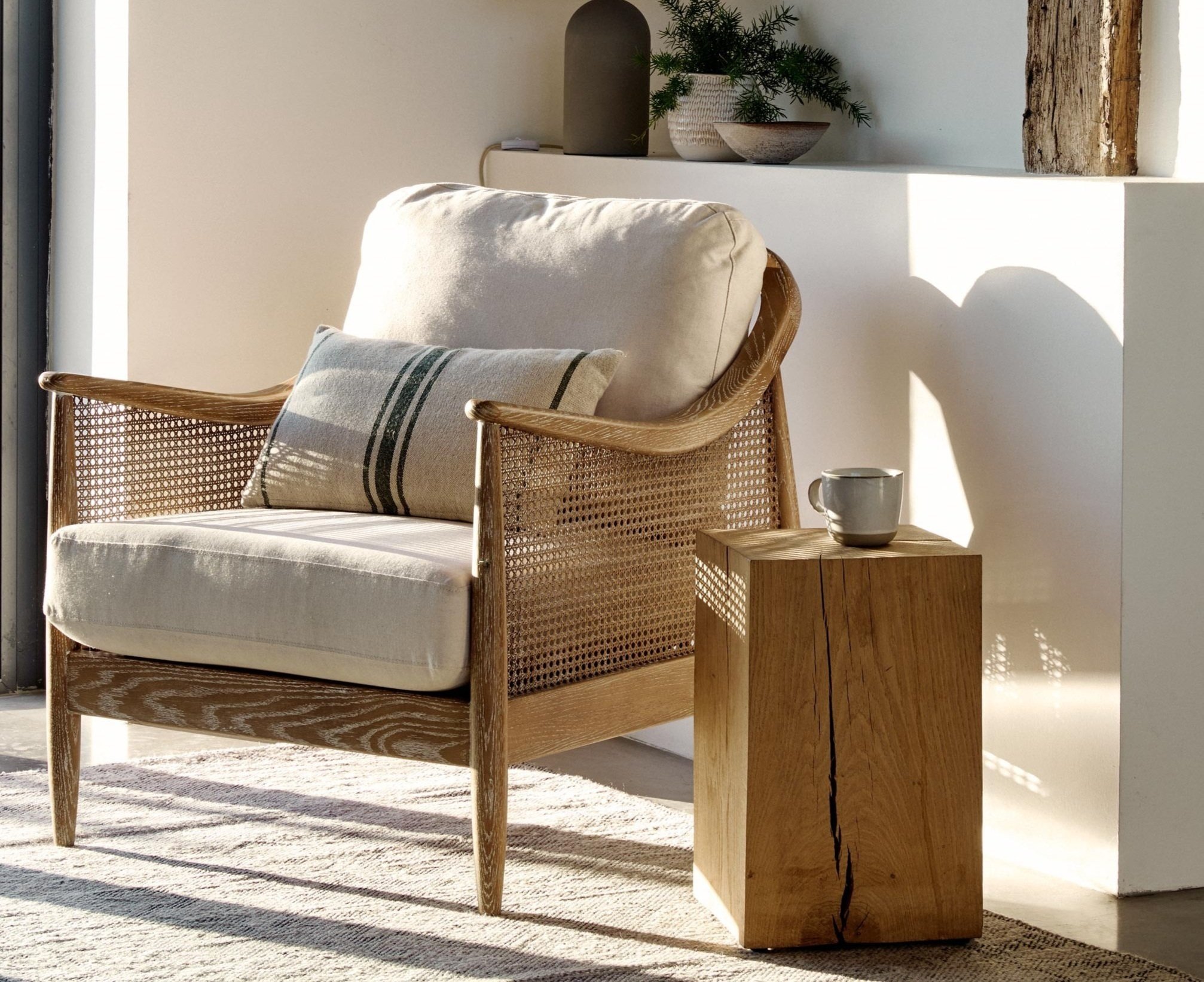 Cozy living space with a woven armchair, beige cushions, and throw pillow. A wooden side table holds a gray mug. Natural light and a neutral color scheme create a serene atmosphere. A small plant and decorative bowl sit on the ledge behind the chair.