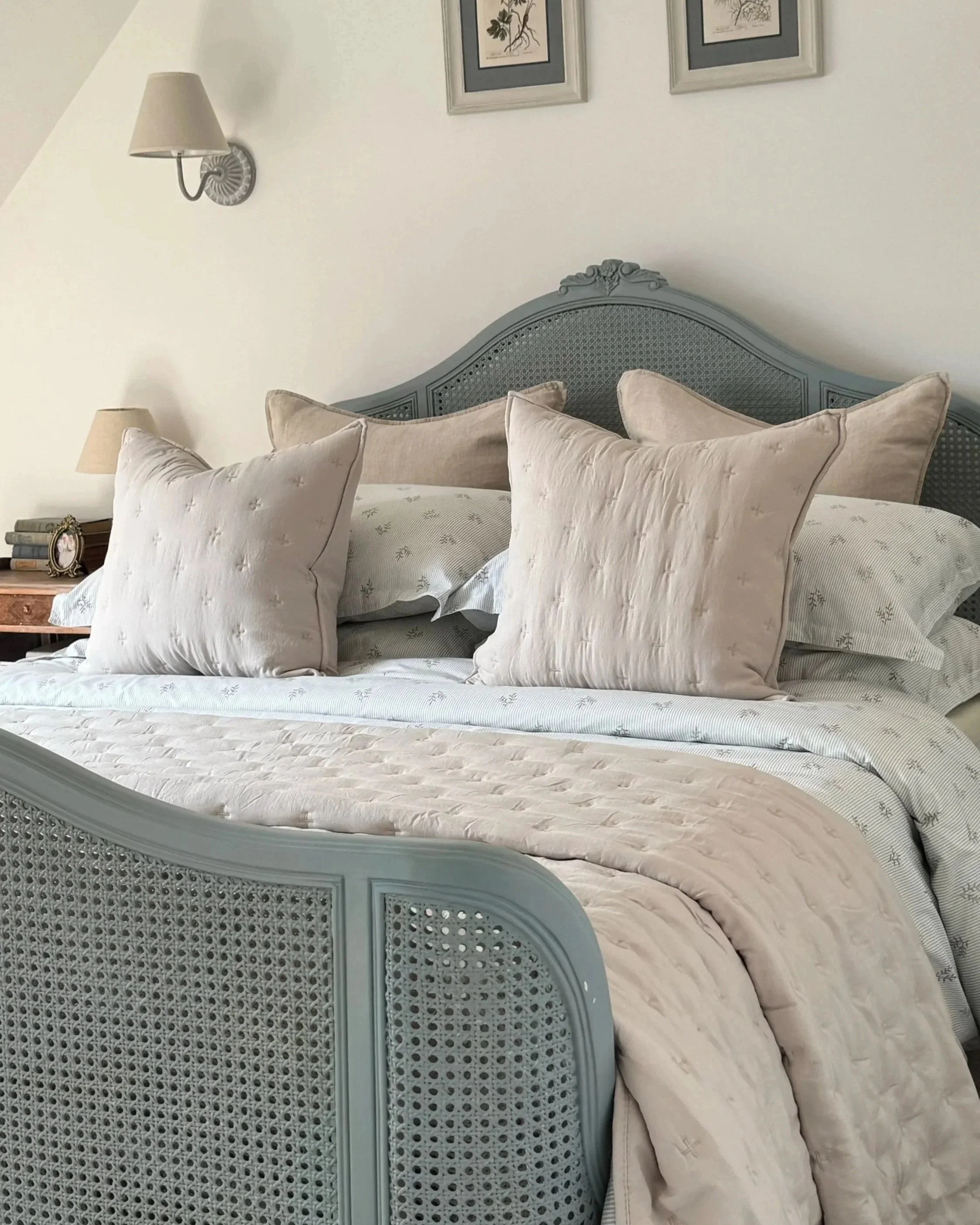 A bedroom with a blue wooden bed frame, soft peachskin bedspread and two cushions. Two wall-mounted lamps and framed botanical prints are above the bed. The side table has books, a picture frame, and a lamp.