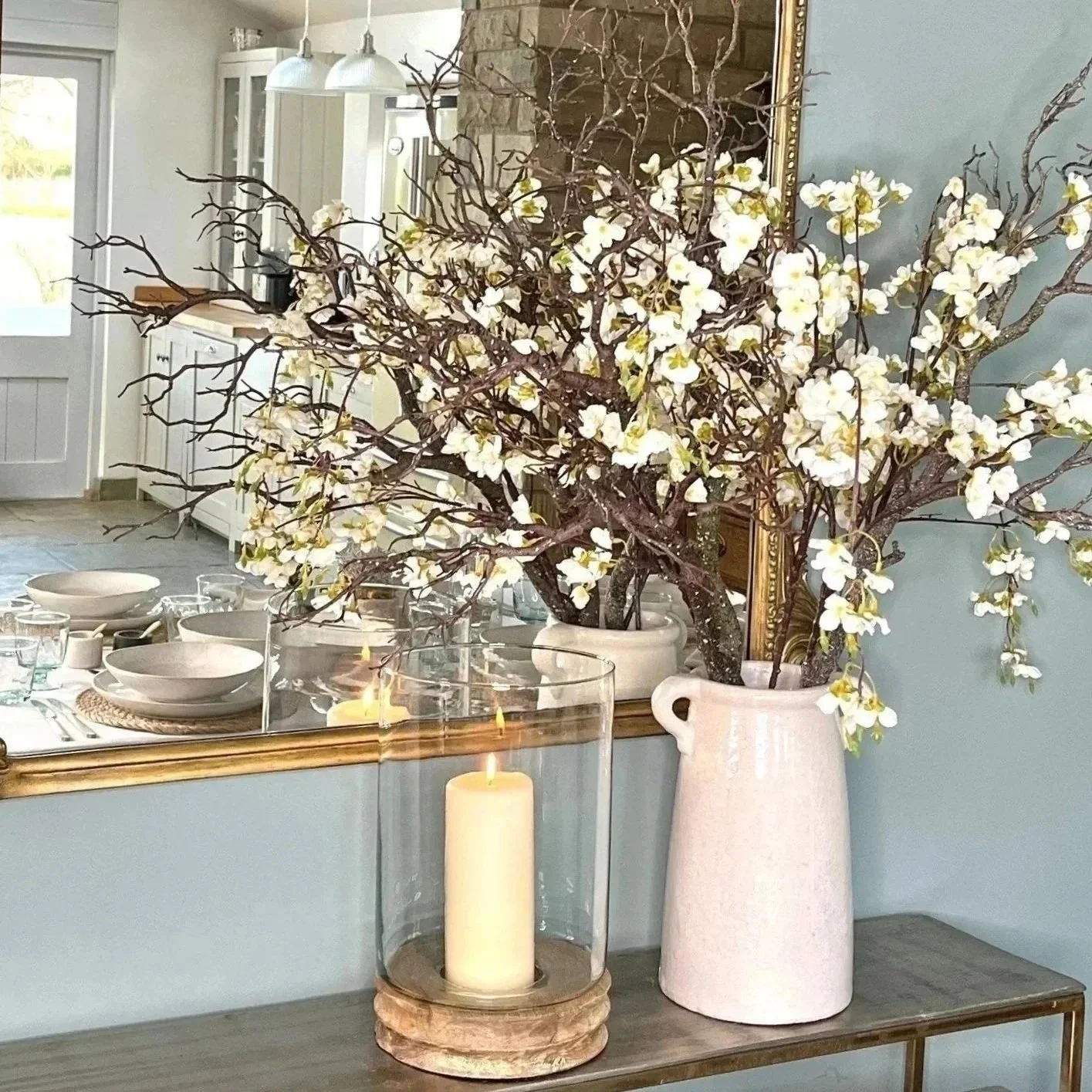 A white ceramic vase with a branch of white flowers and twisted twigs, situated on a wooden side table with a lit candle in a glass hurricane holder in front. Part of a dining room with a table set with plates, bowls, glasses, and silverware, and a living room with a fireplace is visible in the background.