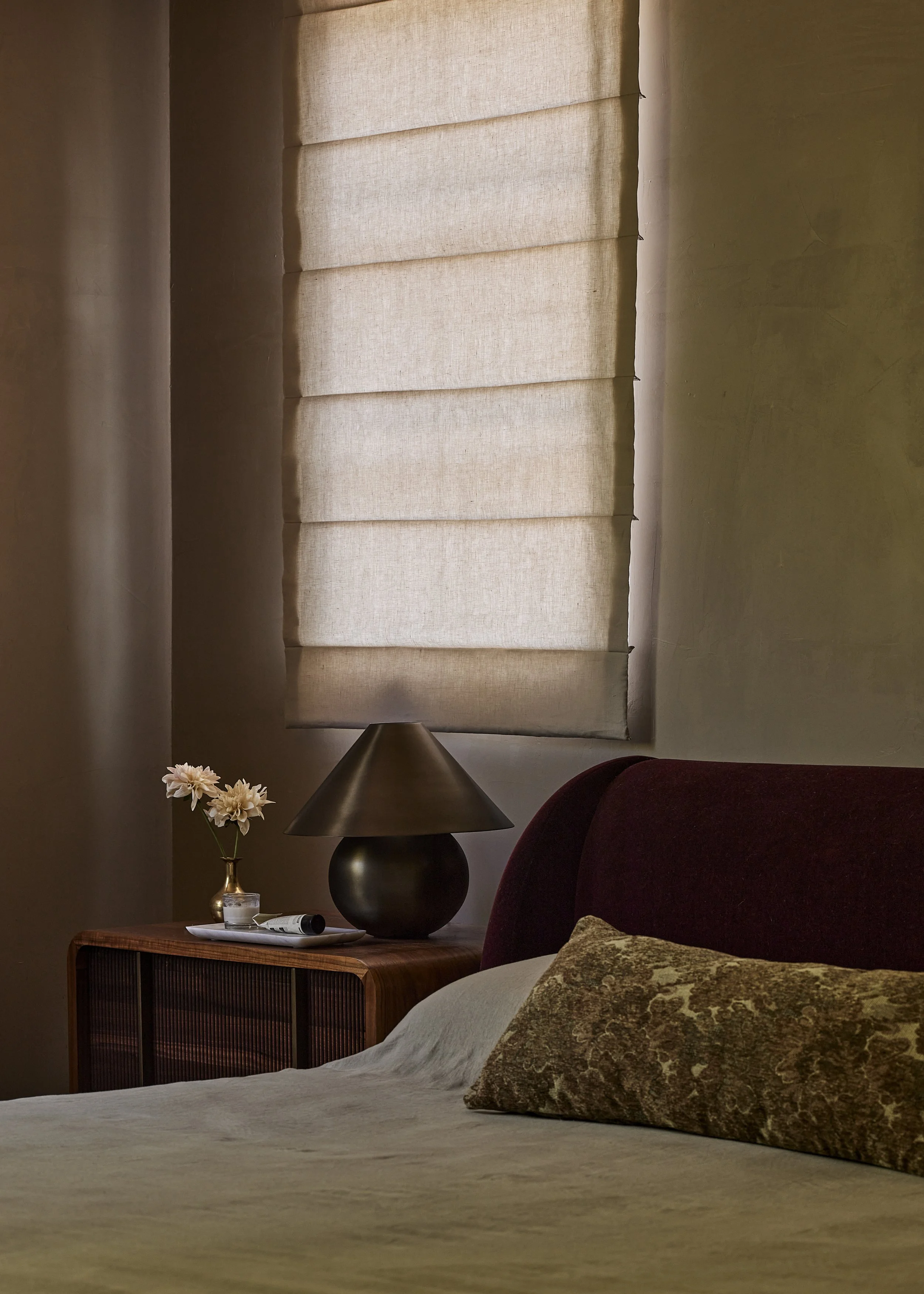 Moody, high-end bedroom vignette in Montecito designed by a Southern California interior designer, featuring a fluted walnut nightstand, sculptural bronze sphere table lamp, marble tray styling, and a deep burgundy velvet upholstered headboard.