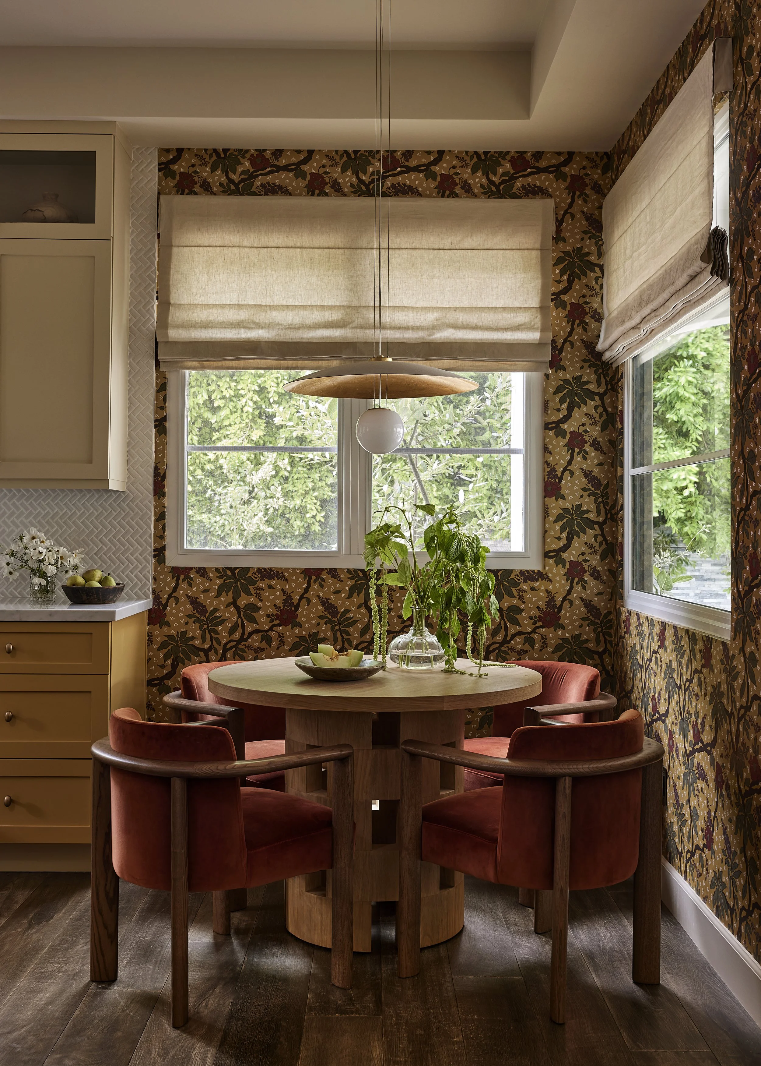 Custom breakfast nook with botanical wallpaper, rust velvet dining chairs, round oak pedestal table, and natural linen Roman shades.