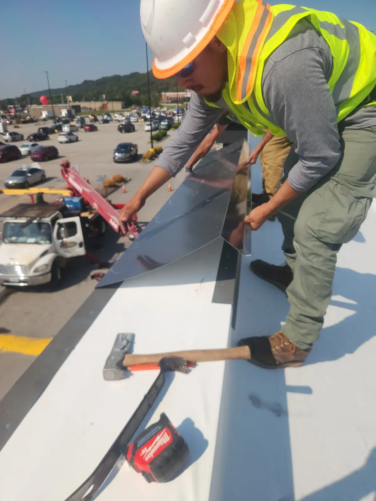 Workers atop a roof installing metal flashing, with construction tools such as a hammer and cordless drill nearby, and a parking lot with cars and a store in the background.