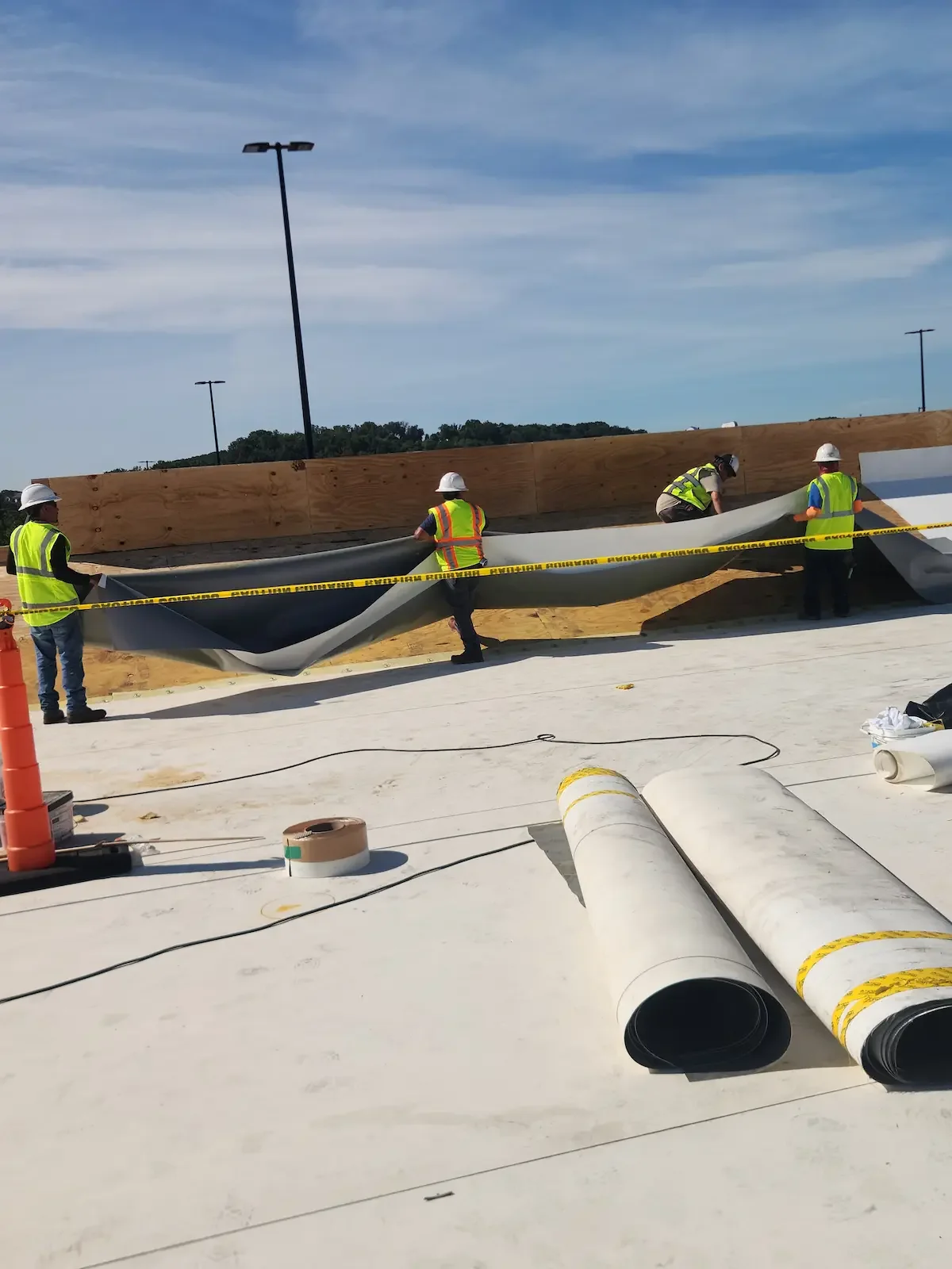 Construction workers installing a large black and gray membrane on a flat surface, with wooden barrier and orange traffic cone nearby, under partly cloudy sky.