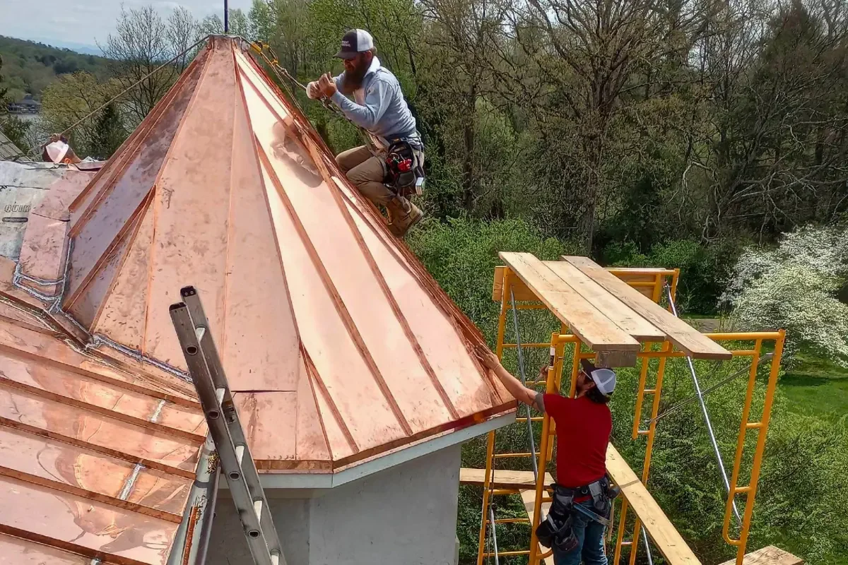 Two construction workers installing a copper roof on a house, with one worker on the roof securing copper panels and the other worker on scaffolding assisting.