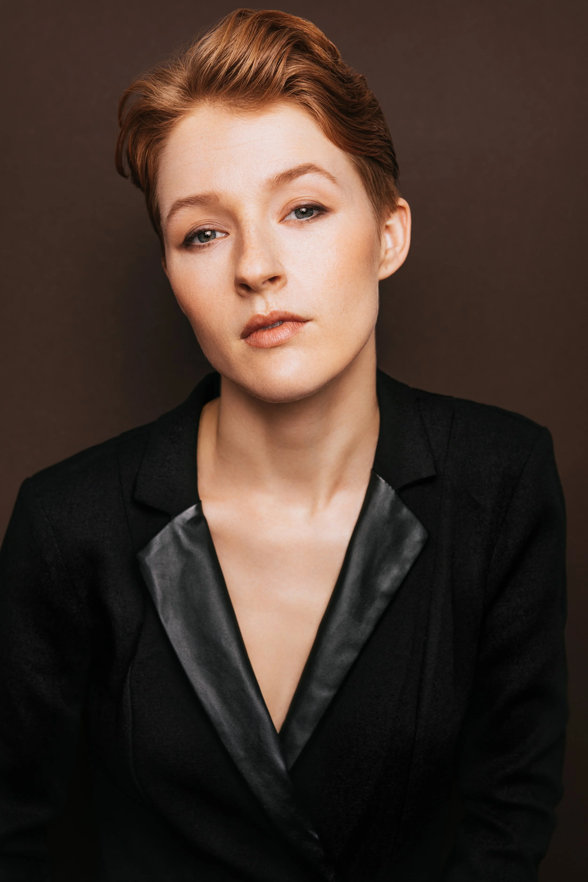 Hawley Gould, in a black blazer with a leather lapel, smirking at camera with chin lifted & tilted, on a dark brown background

by Julianna McGuirl