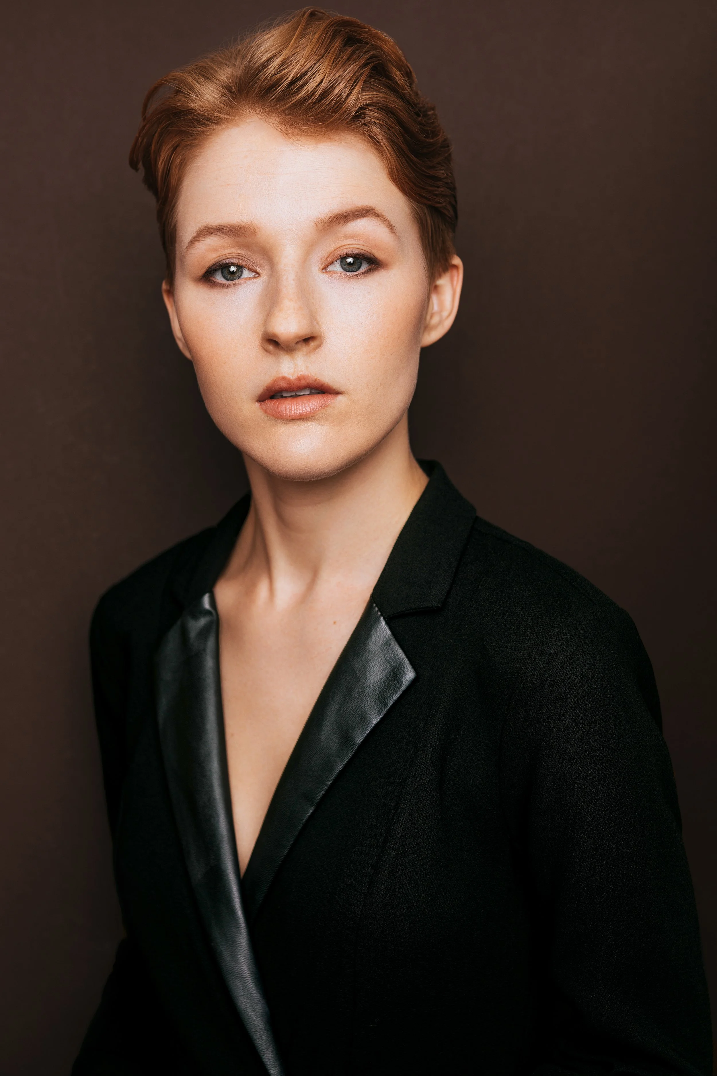 Hawley Gould in a blazer with leather lapel, smokey eyeliner and a strong pose, looking right down the barrel of the camera, on a dark brown background

by Julianna McGuirl