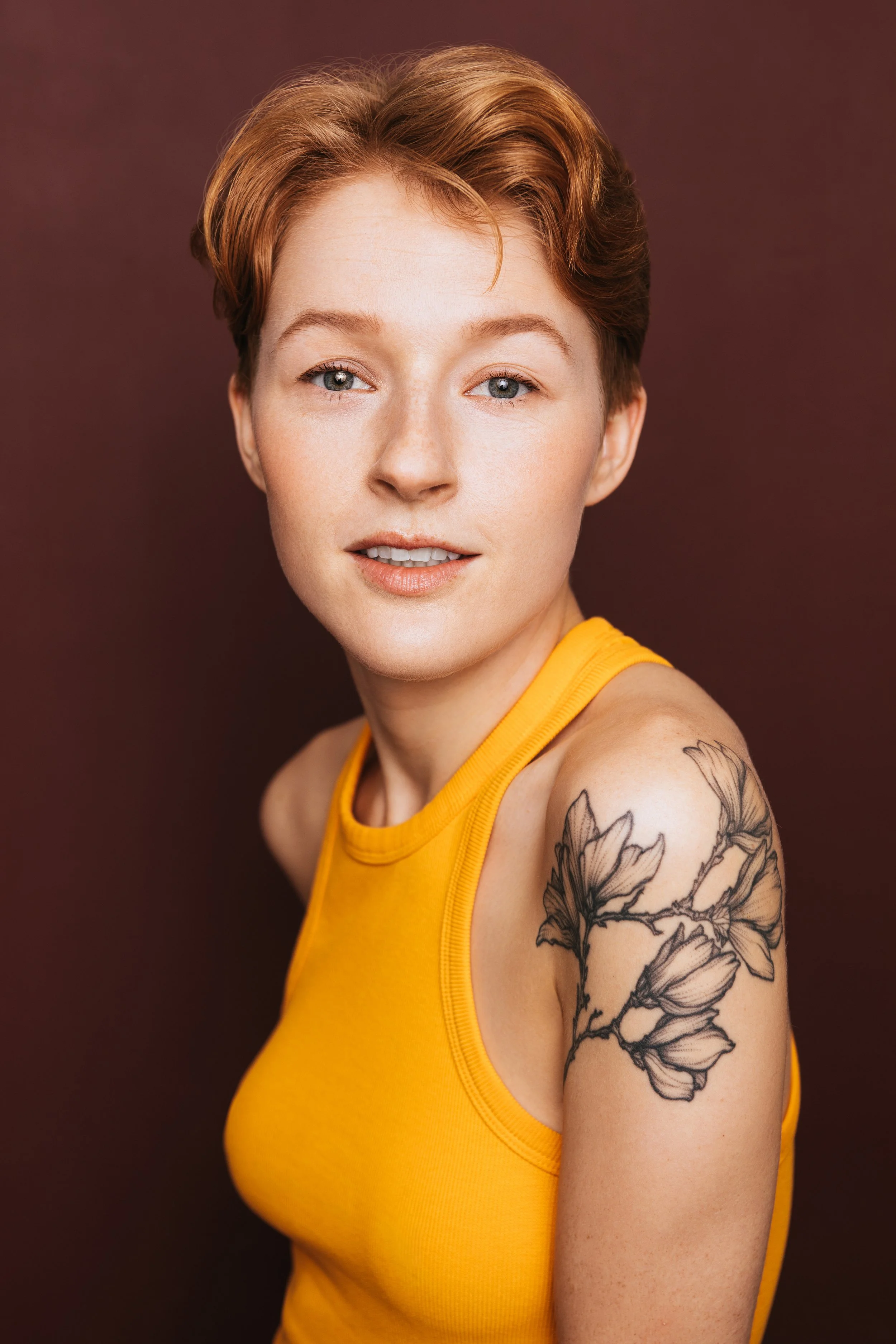 Hawley Gould in side profile with face towards camera, in an orange tank, magnolia tattoo visible on shoulder, half smiling knowingly, on a burgundy background

by Julianna McGuirl 
