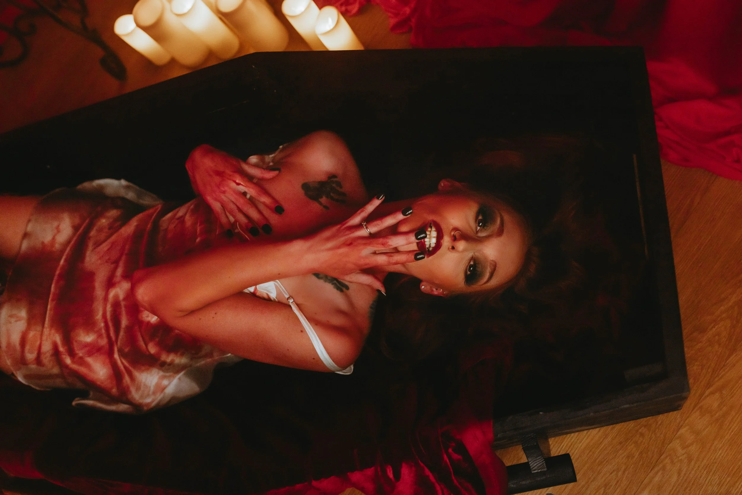 A woman with tattoos and dark makeup lying on a black surface, wearing a revealing satin dress, covering her mouth with her hand, surrounded by candles and red satin fabric.