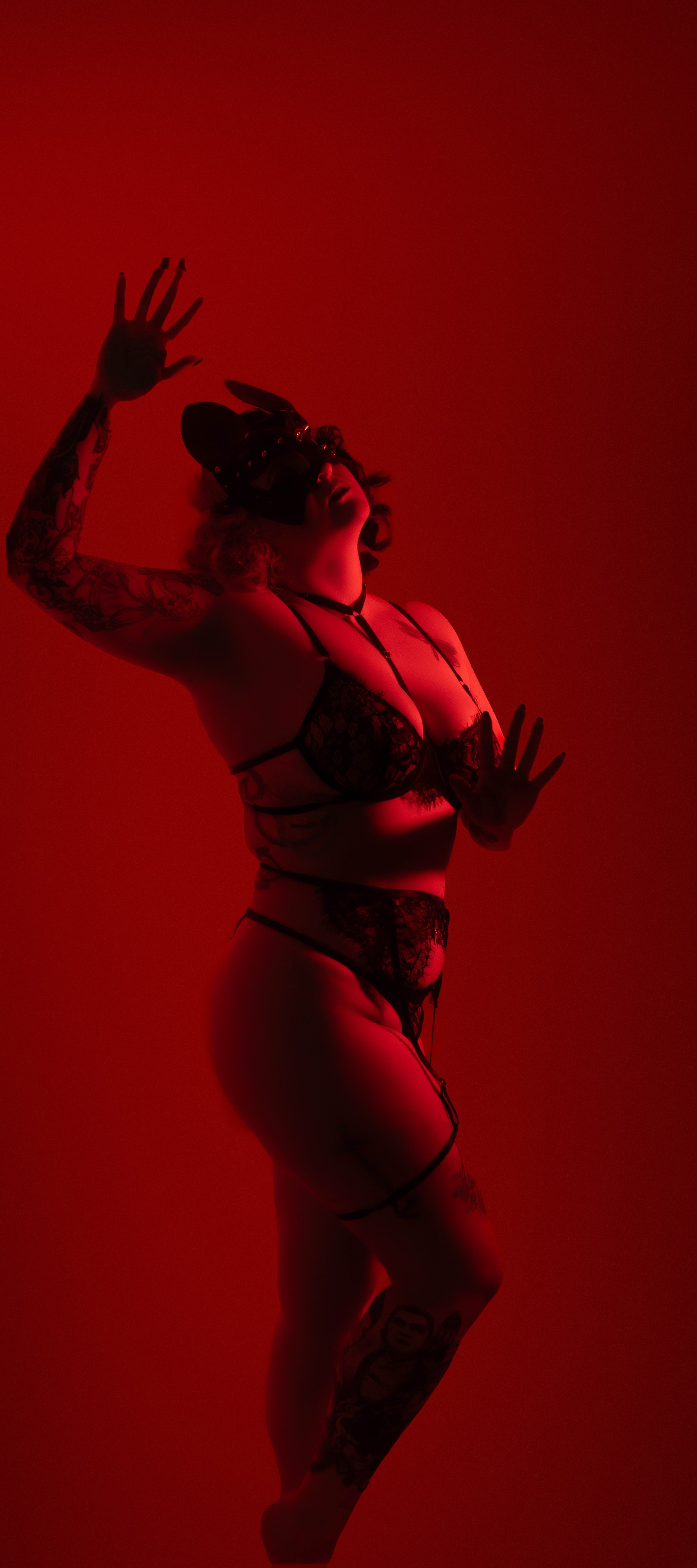 Woman in black lace lingerie, wearing bunny ears mask, posing against a red background with dramatic lighting.