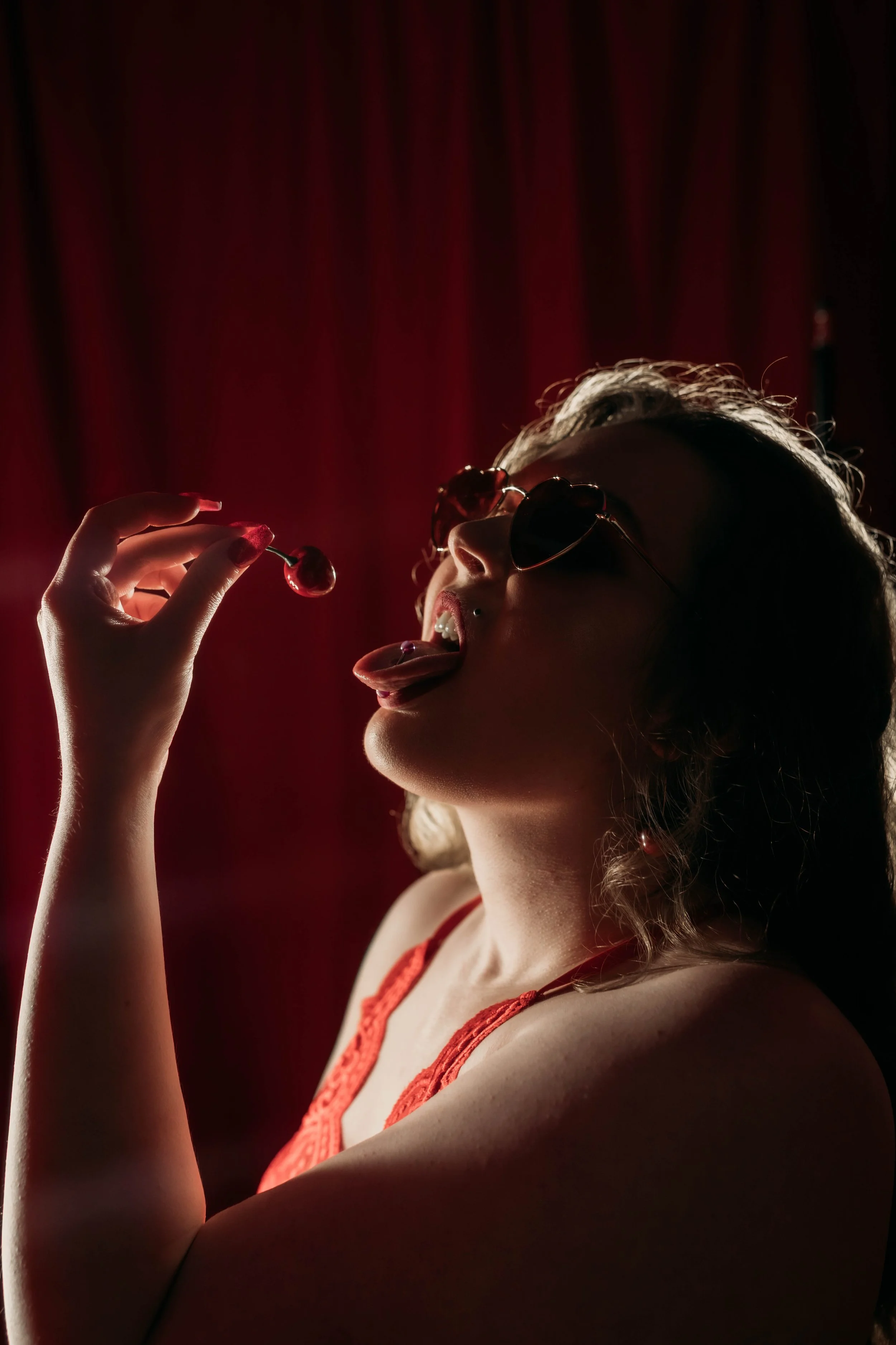 Young woman with curly hair, wearing sunglasses and an orange top, holding a cherry near her mouth and licking her tongue out, set against a red curtain background.