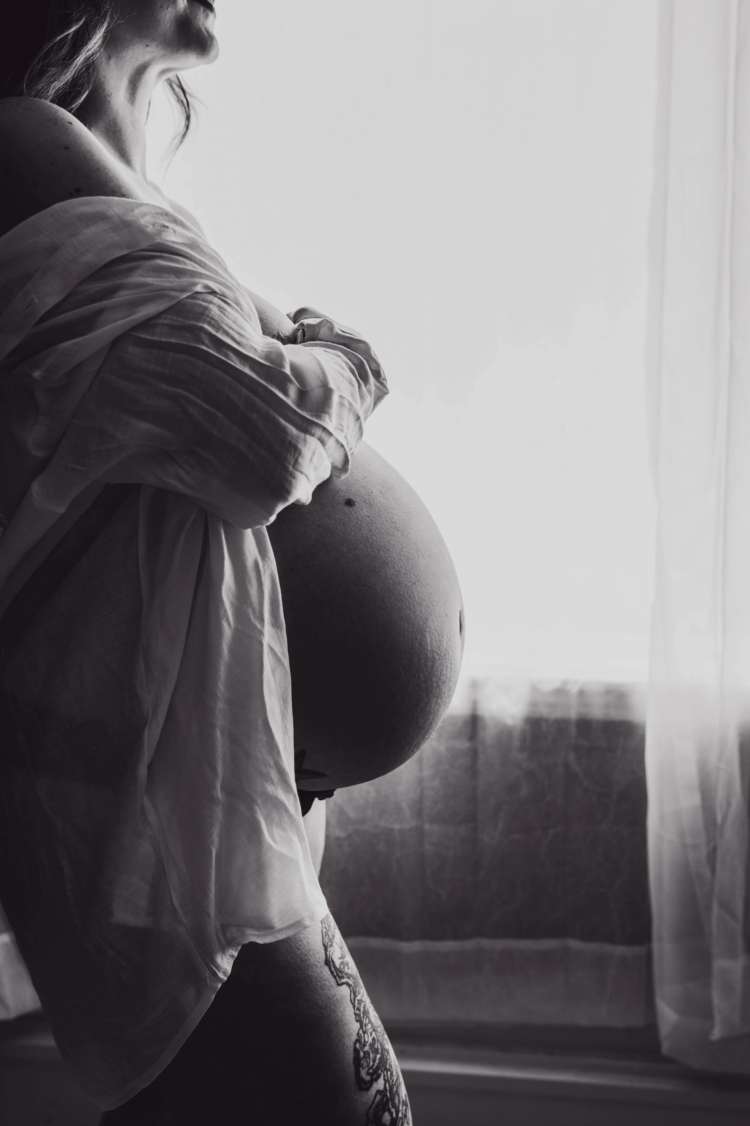 Black and white photo of a pregnant woman standing by a window, cradling her belly with both hands, wearing a loose shirt, with a tattoo on her thigh.