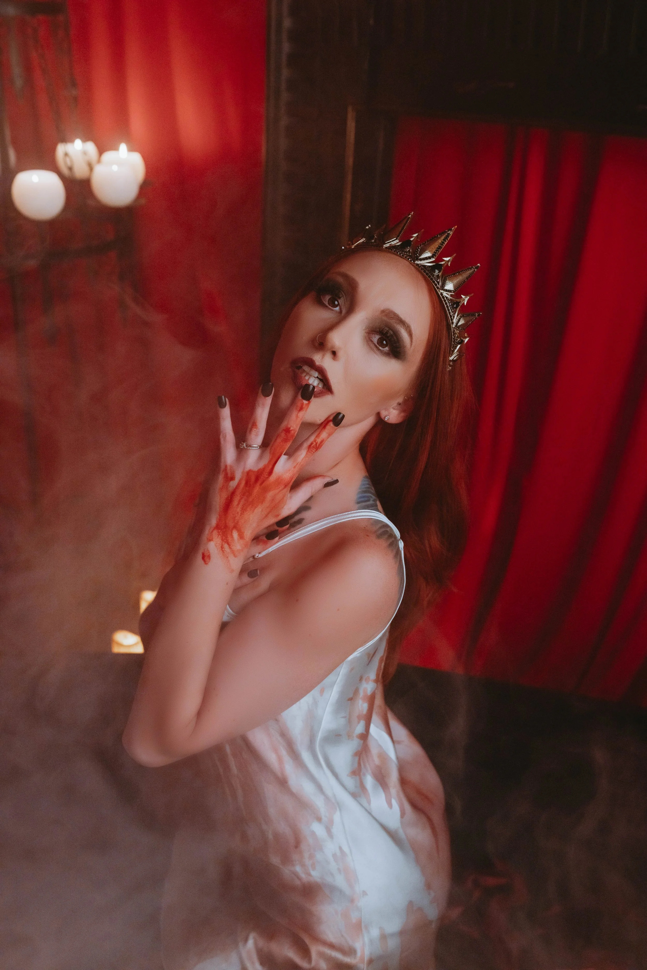A woman with red hair wearing a crown, makeup, and a white satin dress, holding her hand with blood-like makeup around her fingers, in a dimly lit room with red curtains and candles, with smoke or fog around her.