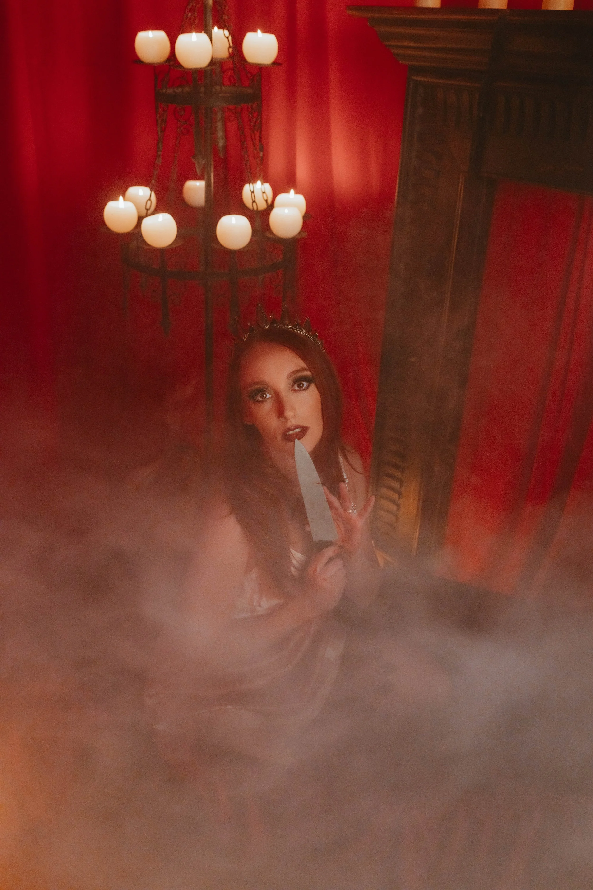 A woman dressed as a witch with dark makeup and a crown, holding a kitchen knife, surrounded by fog in a red room with candle chandelier and a wooden cabinet.