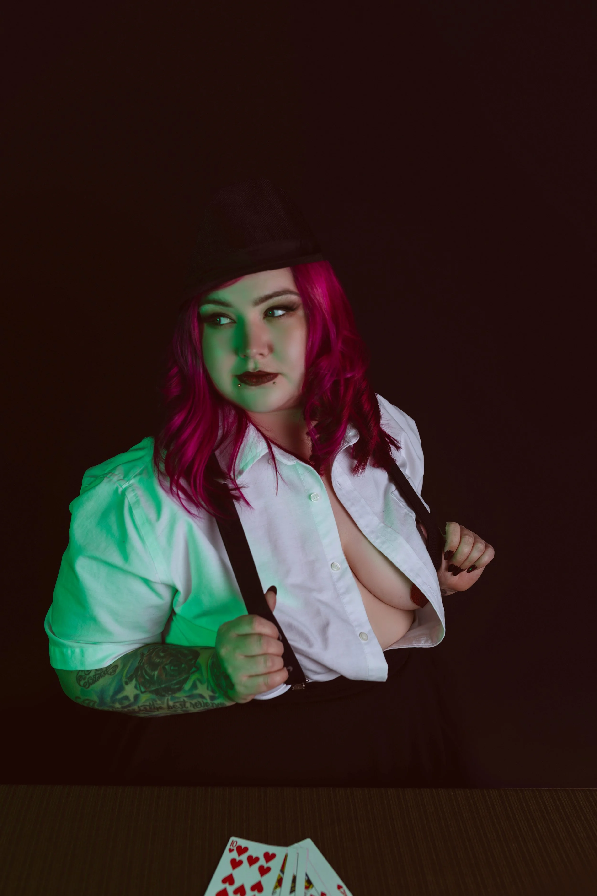 A woman with bright pink hair, wearing a black hat and white shirt, partially unbuttoned, sitting at a table with playing cards, with a black background and green/red lighting effects.