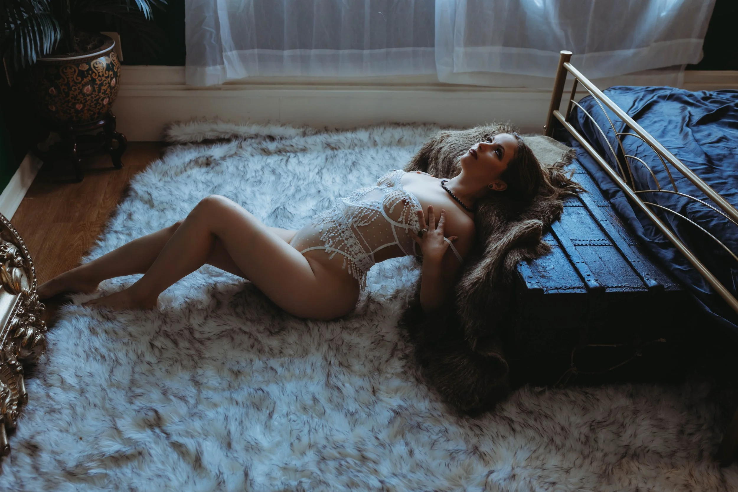 Young woman lying on a plush white rug beside a bed, wearing a white lace lingerie, with her head resting on a faux fur blanket, in a warmly lit bedroom with sheer curtains.