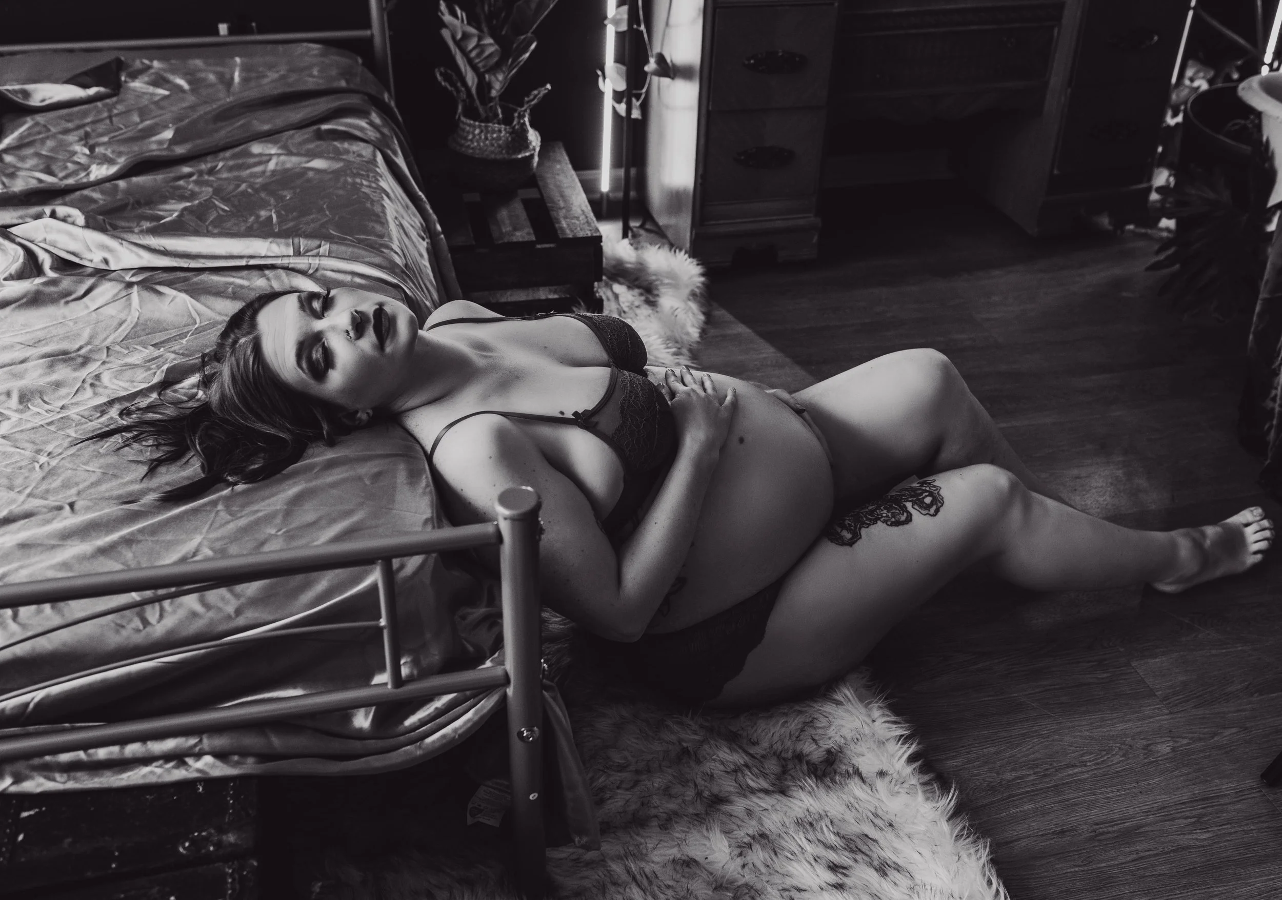 A woman with long hair lying on a rug beside a bed wearing black lingerie, with her hand on her stomach, in a bedroom with wooden furniture and a potted plant, in black and white.