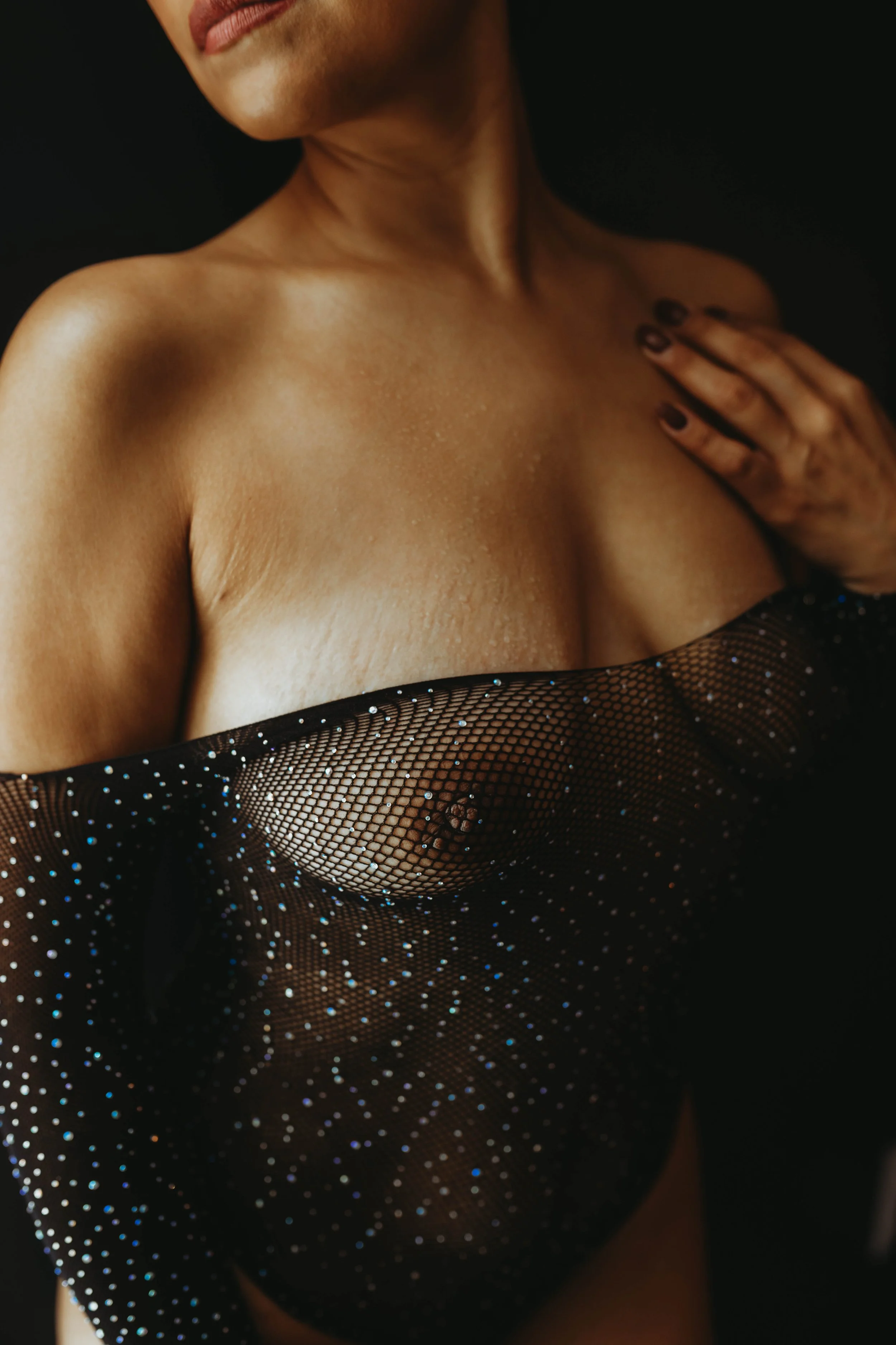 Close-up of a woman with dark skin, wearing a sheer black garment with rhinestones, covering her chest with her left hand touching her collarbone.