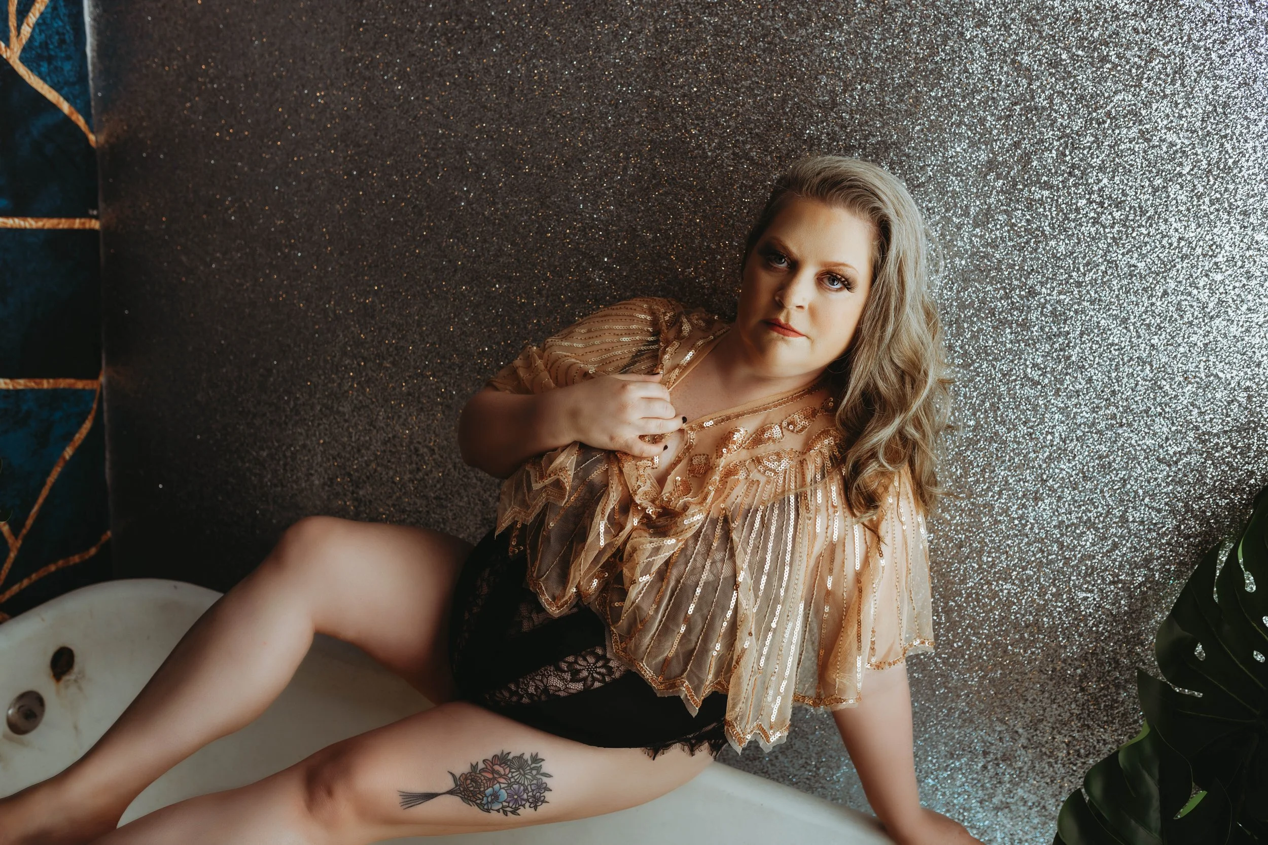 A woman with wavy blonde hair and a tattoo of flowers on her thigh, sitting in a bathtub against a glittery silver wall, wearing a sheer striped gold top with ruffles.