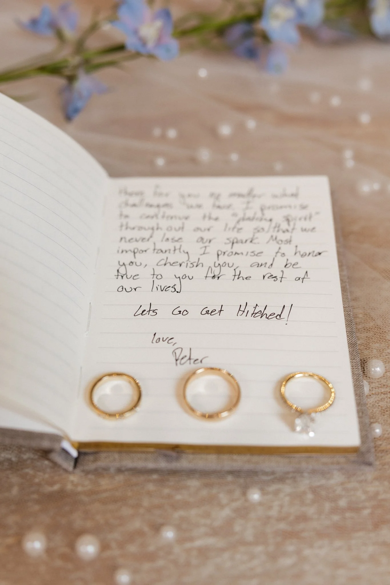 Open notebook with handwritten note and three gold rings on a wooden surface with scattered pearl-like beads and a sprig of blue flowers in the background.