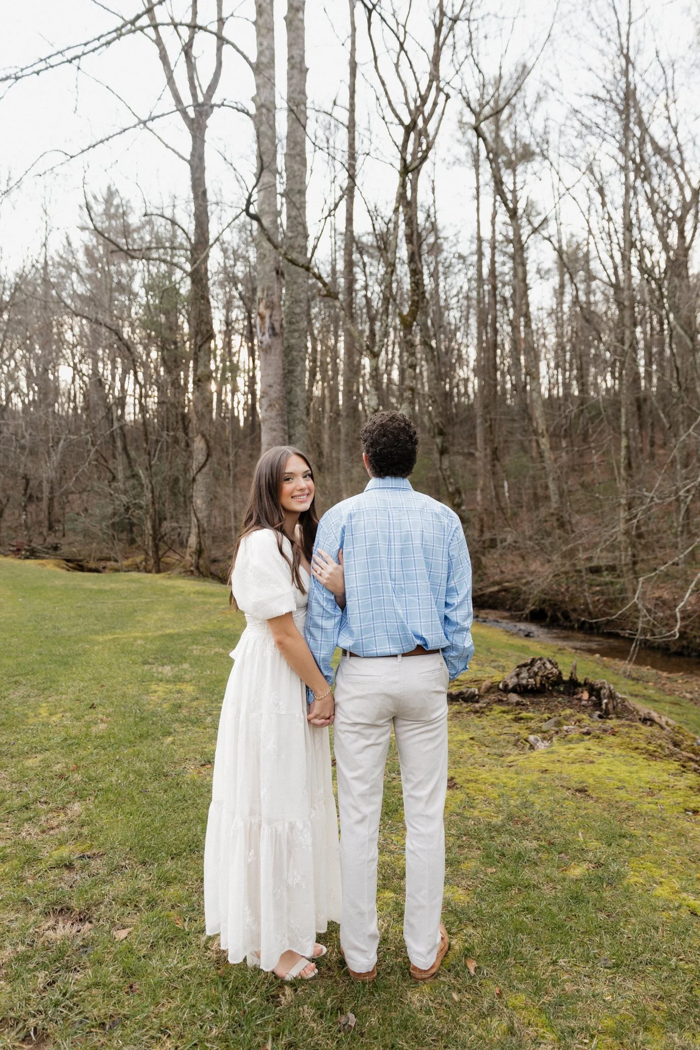 A young couple holding hands outdoors in a grassy area with bare trees in the background, during what appears to be early spring or late autumn.
