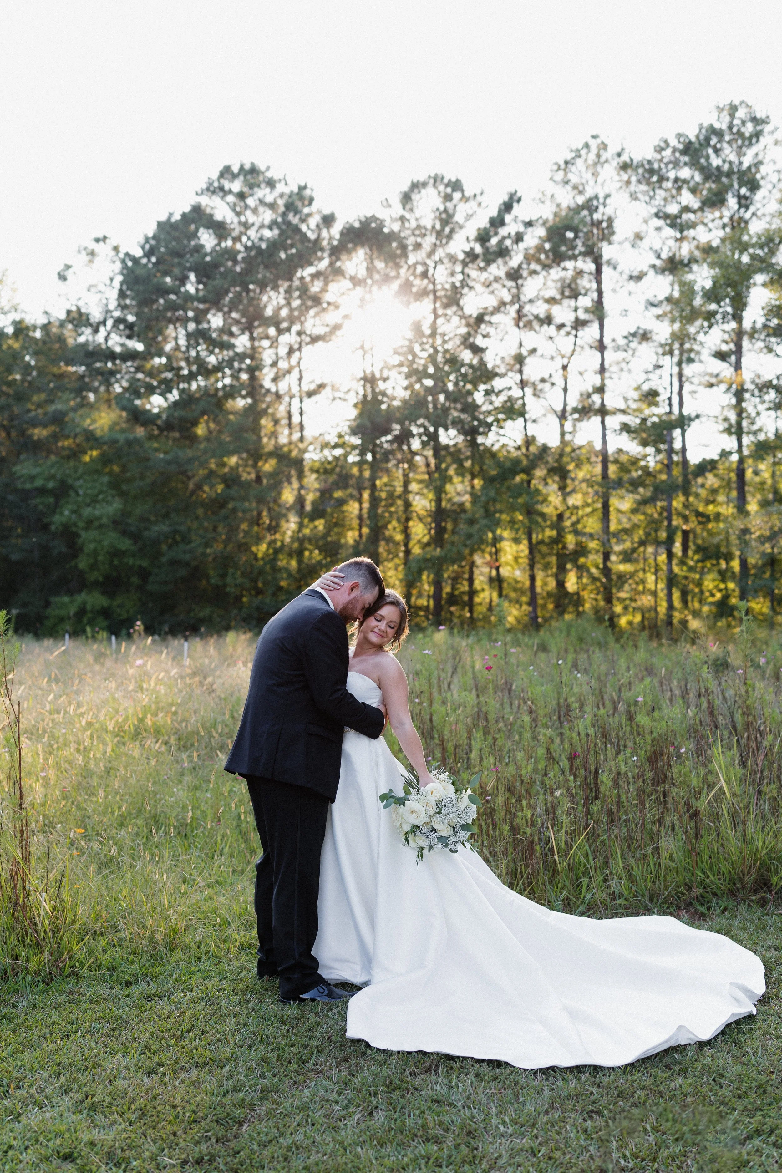 A bride and groom sharing a tender moment outdoors during their wedding, with the bride holding a bouquet of white flowers and greenery, standing in a grassy field with trees and sunlight in the background.