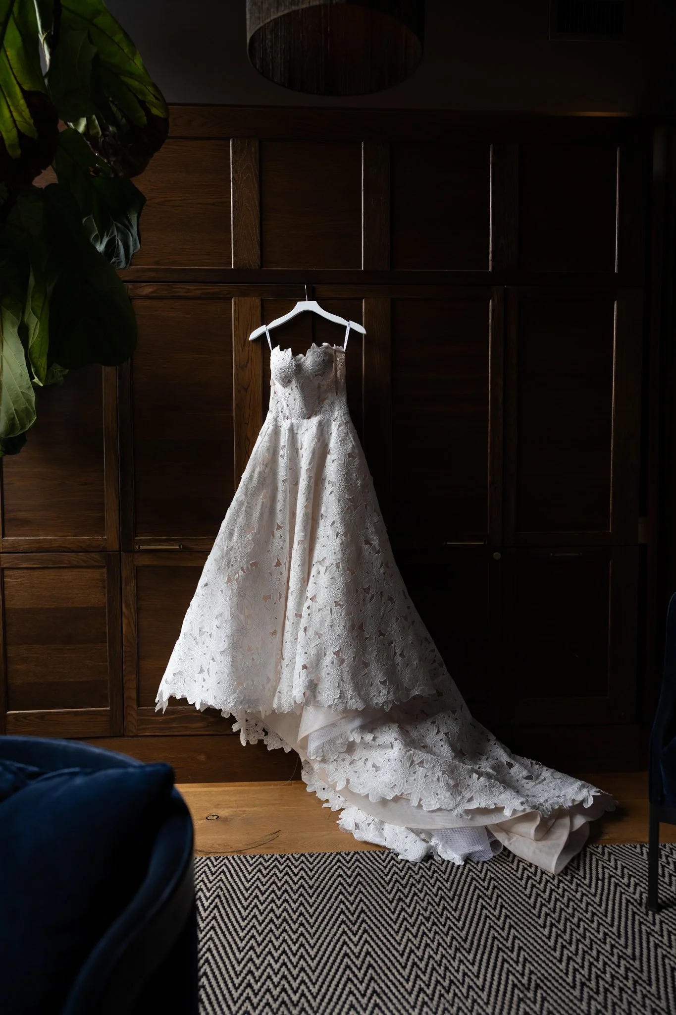A white lace wedding dress hanging on a white hanger against a dark wooden wall.