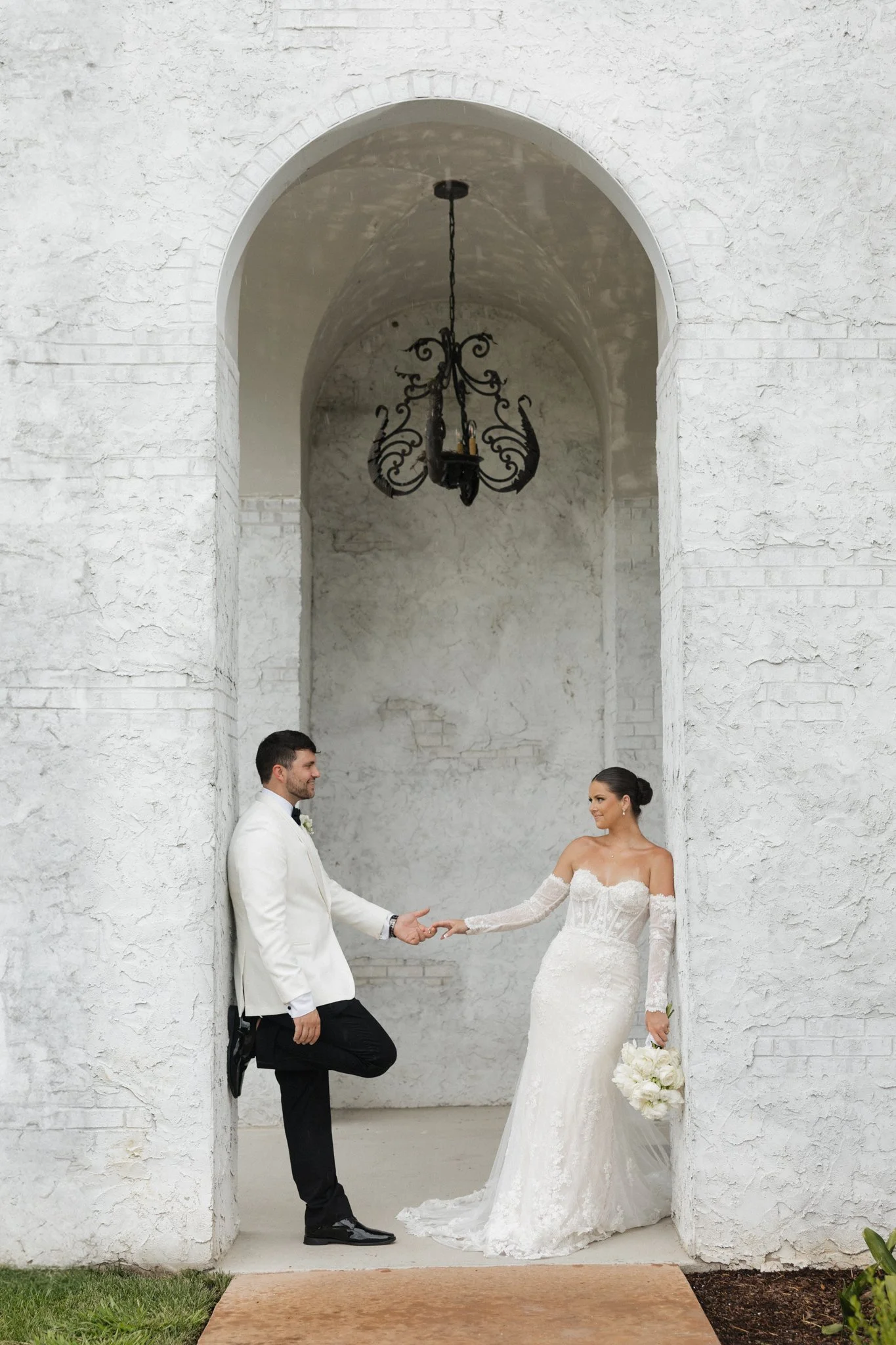 Bride and groom holding hands in front of white textured wall with archway and black chandelier
