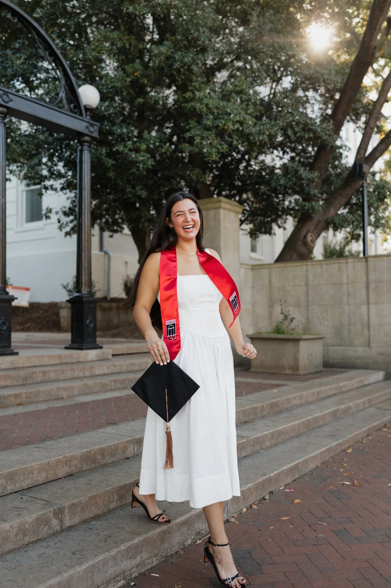 A young woman in a white dress, black high heels, and a red graduation stole holding a black graduation cap, smiling as she finishes her graduation ceremony outdoors under the trees.