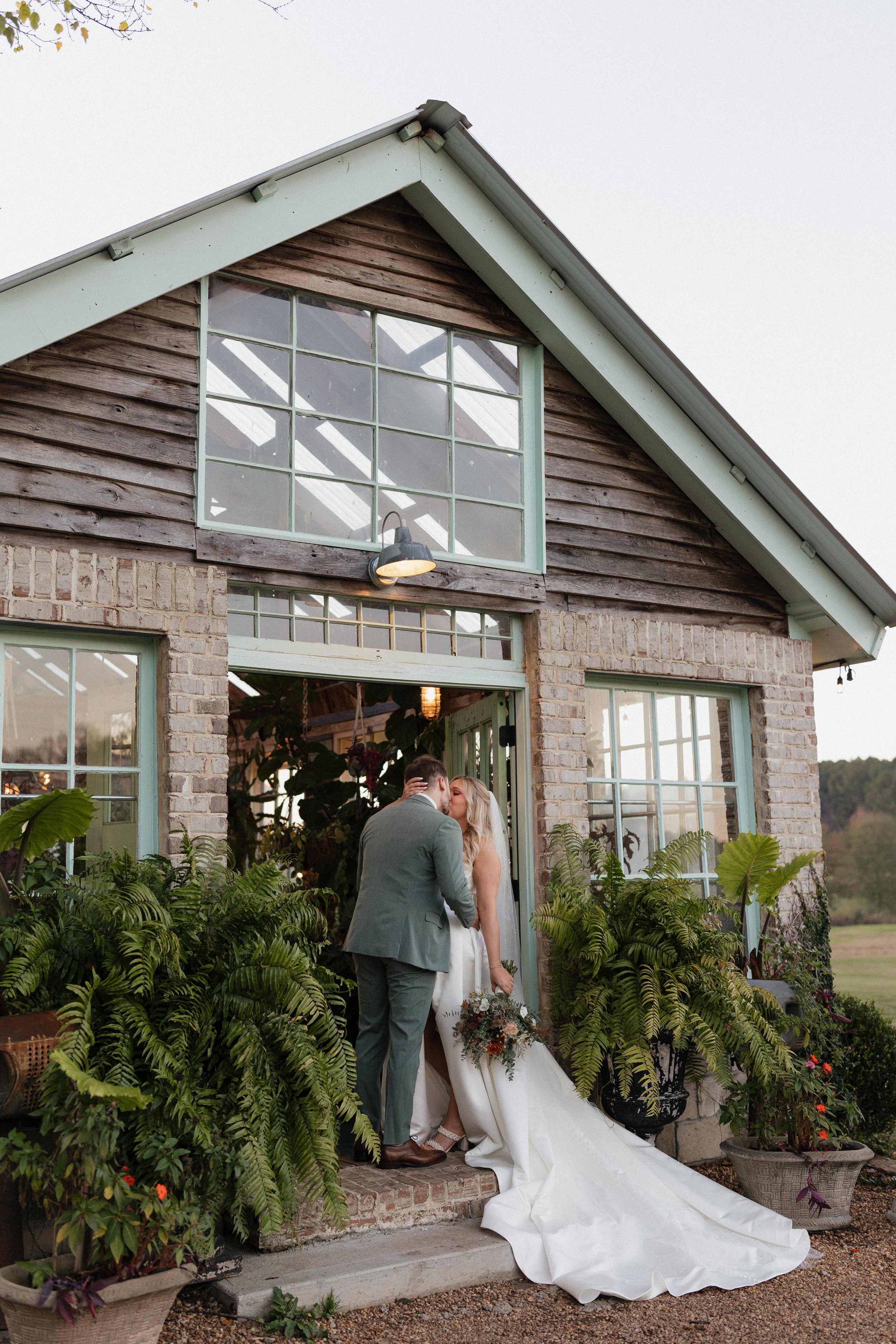 A bride and groom kissing at the entrance of a rustic brick and wood greenhouse surrounded by lush green plants.