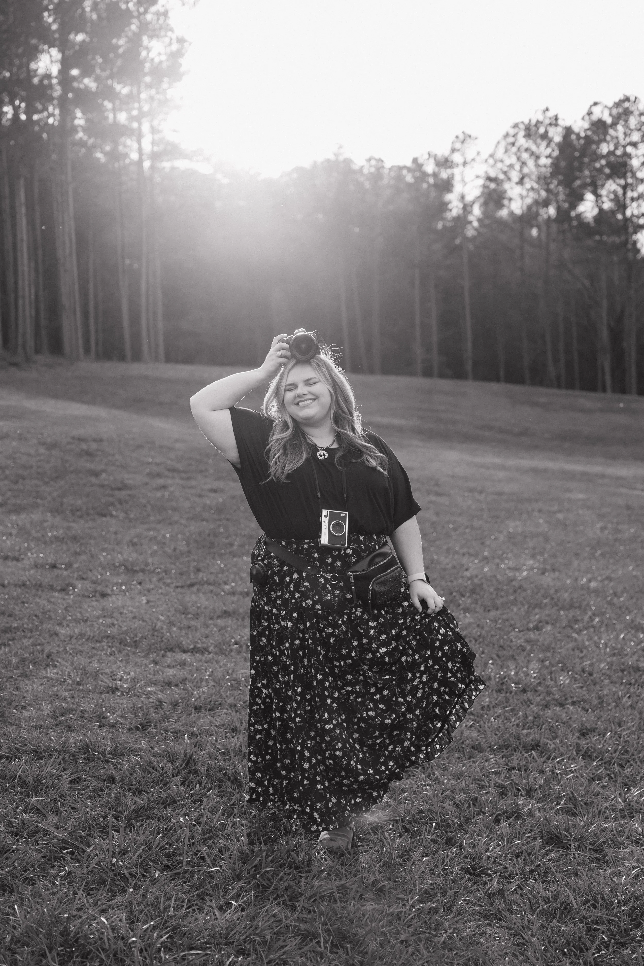 A woman smiling and holding a camera above her head in a grassy outdoor area with trees in the background.