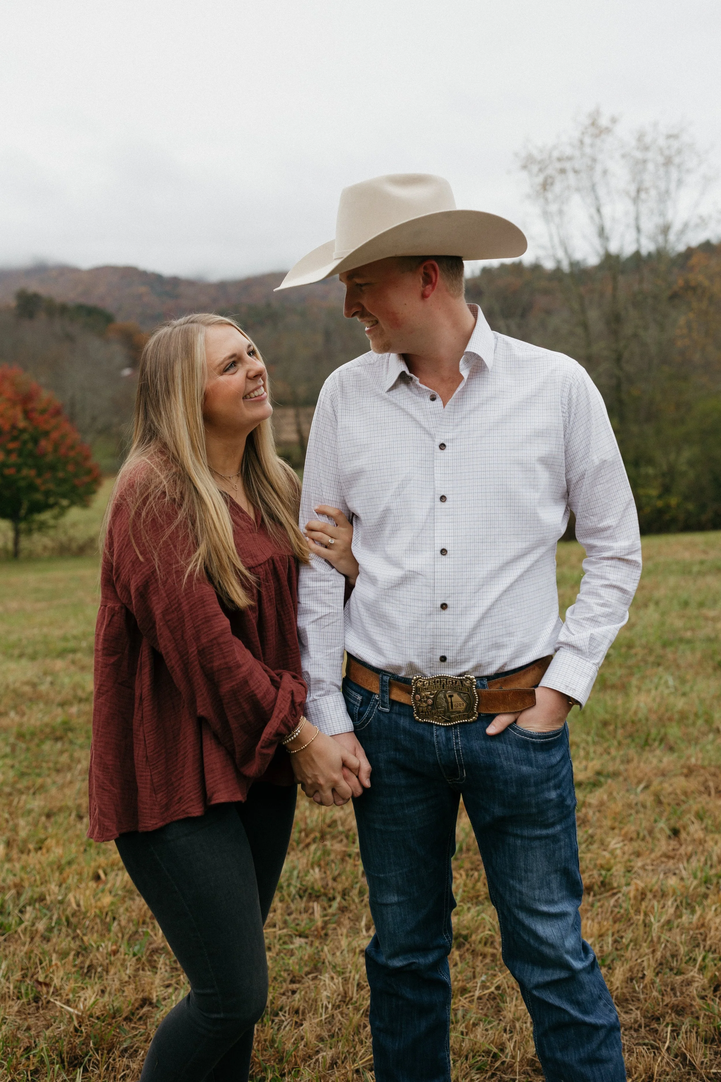 A smiling young couple holding hands outdoors with a man wearing a large cowboy hat and a woman with long blonde hair, in a field with trees and mountains in the background.