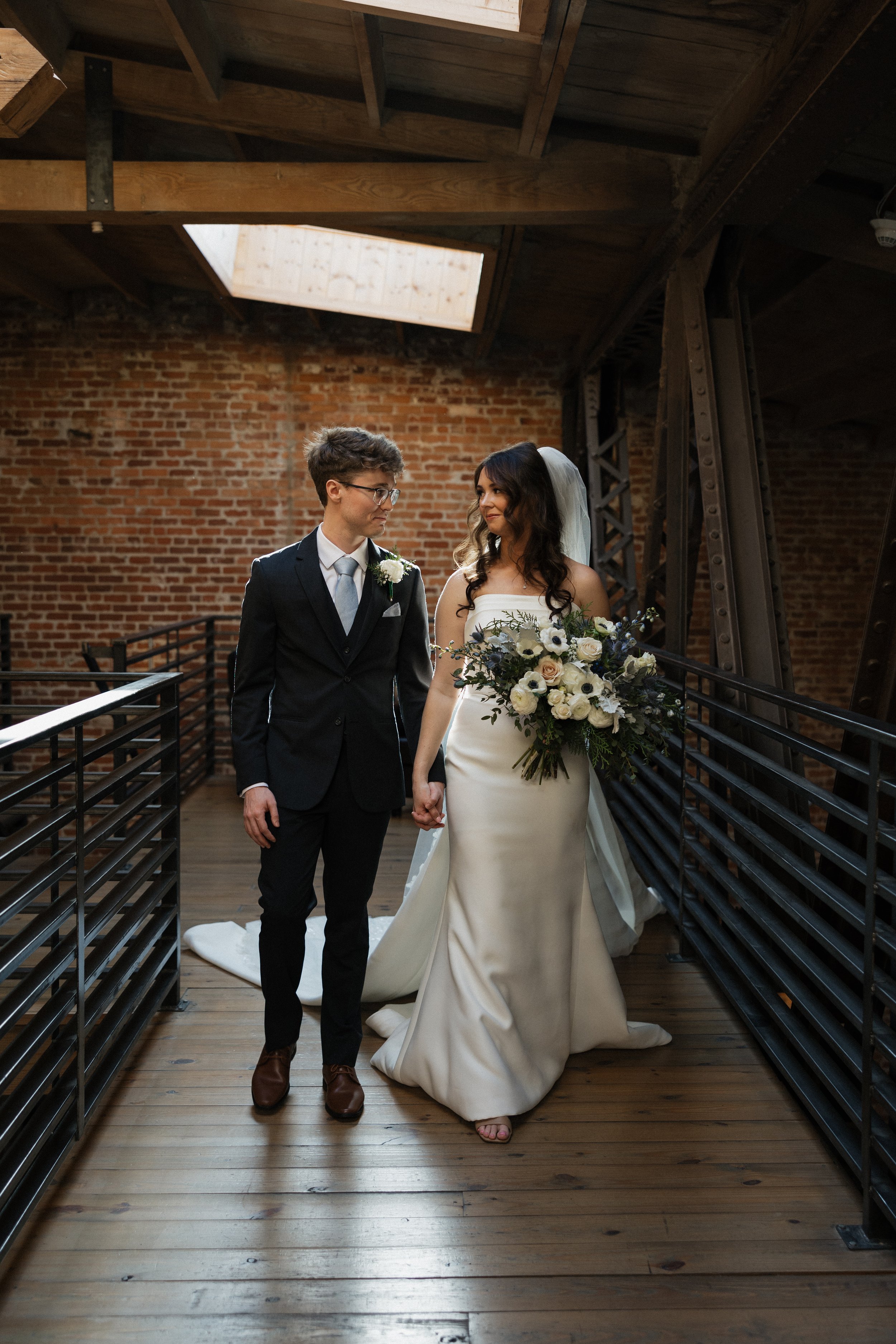 A bride and groom holding hands on a wooden floor in a rustic indoor setting with brick walls and exposed wooden beams. The bride wears a white satin gown and veil, holding a large bouquet of white and dark flowers. The groom wears a black suit, whit