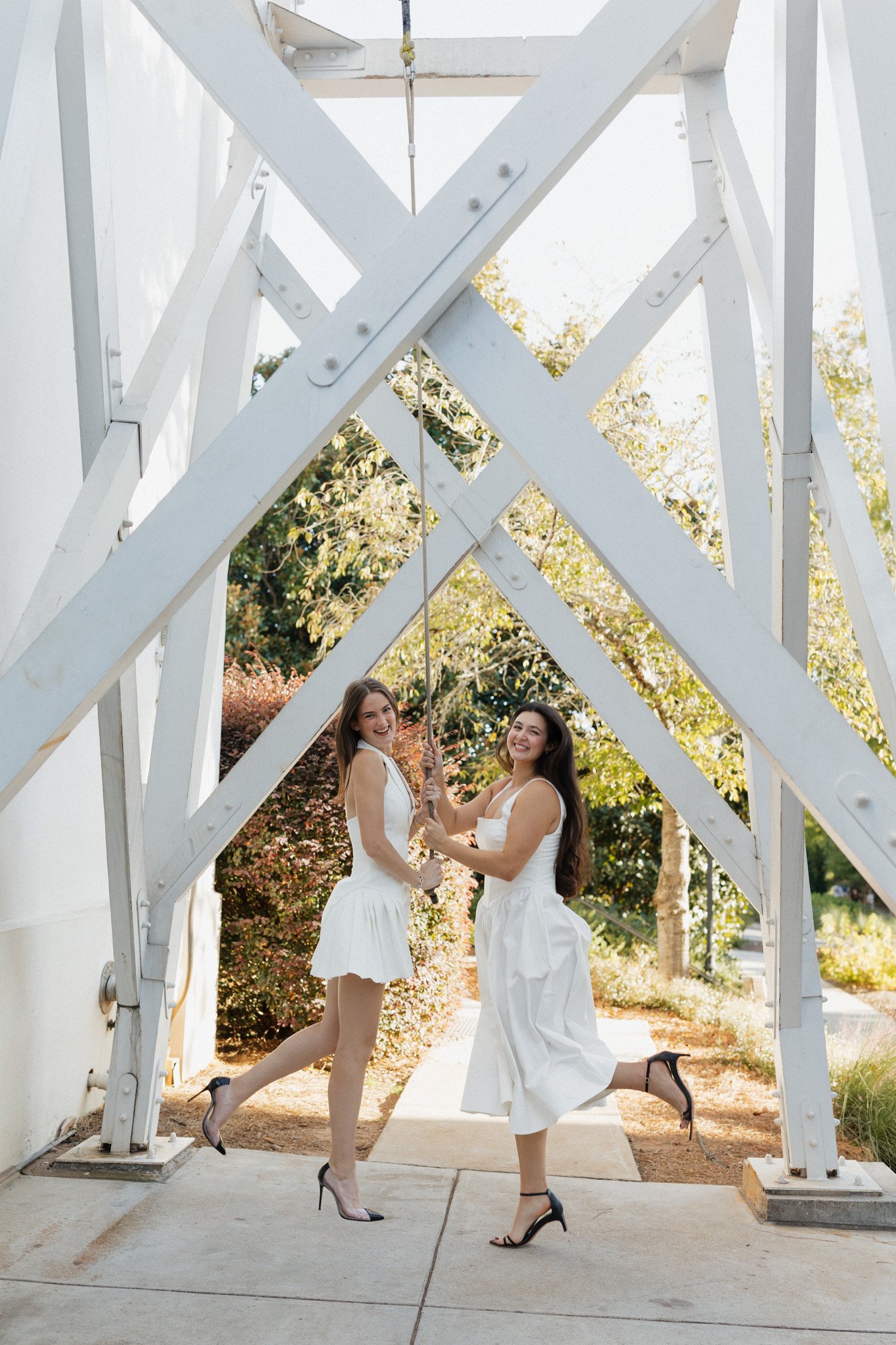 Two women in white dresses holding onto a swing chain underneath a white wooden structure, smiling and posing outdoors with trees and bushes in the background.