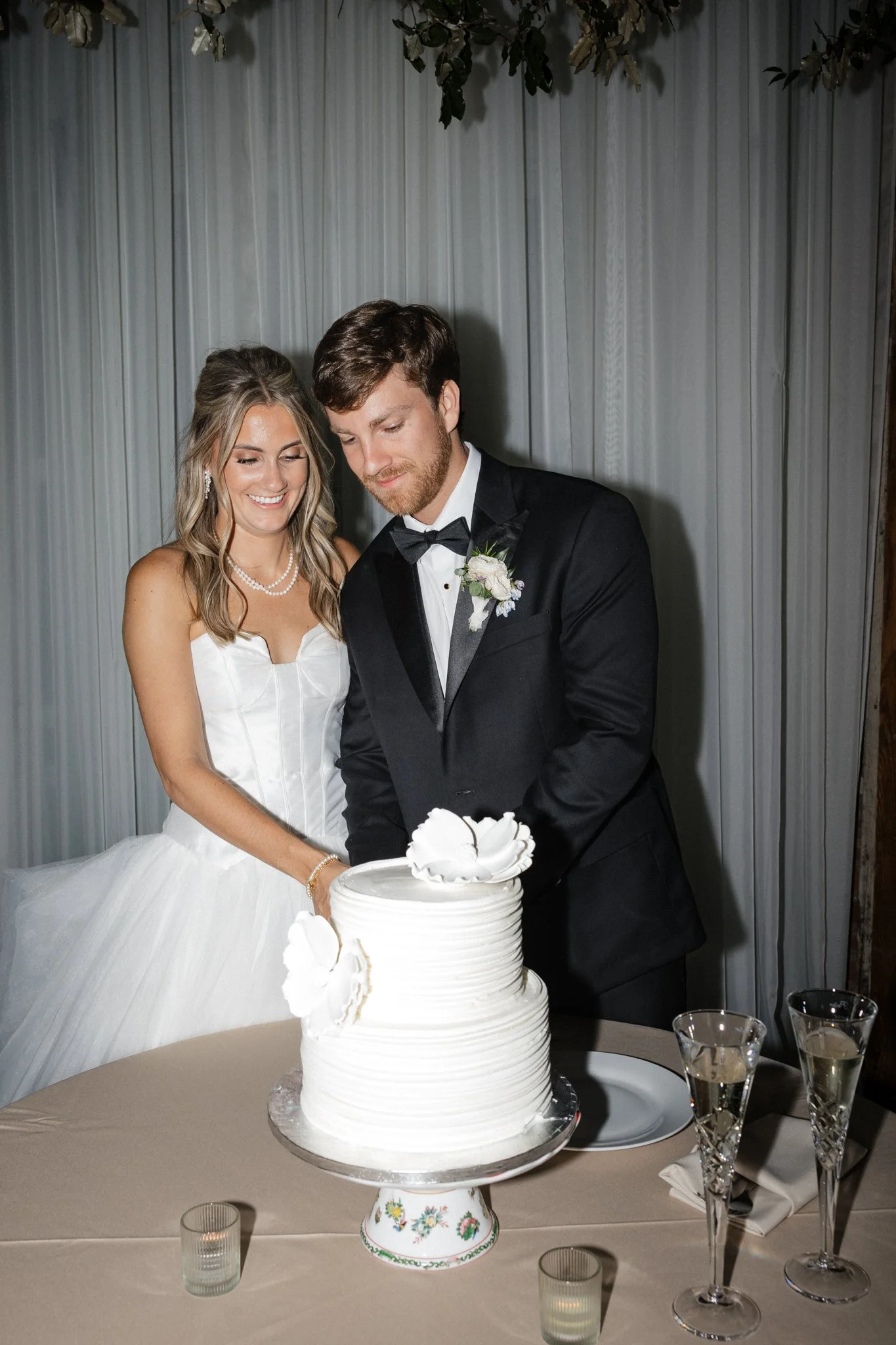 A bride and groom cutting a white wedding cake together during their wedding reception, with champagne glasses on the table.