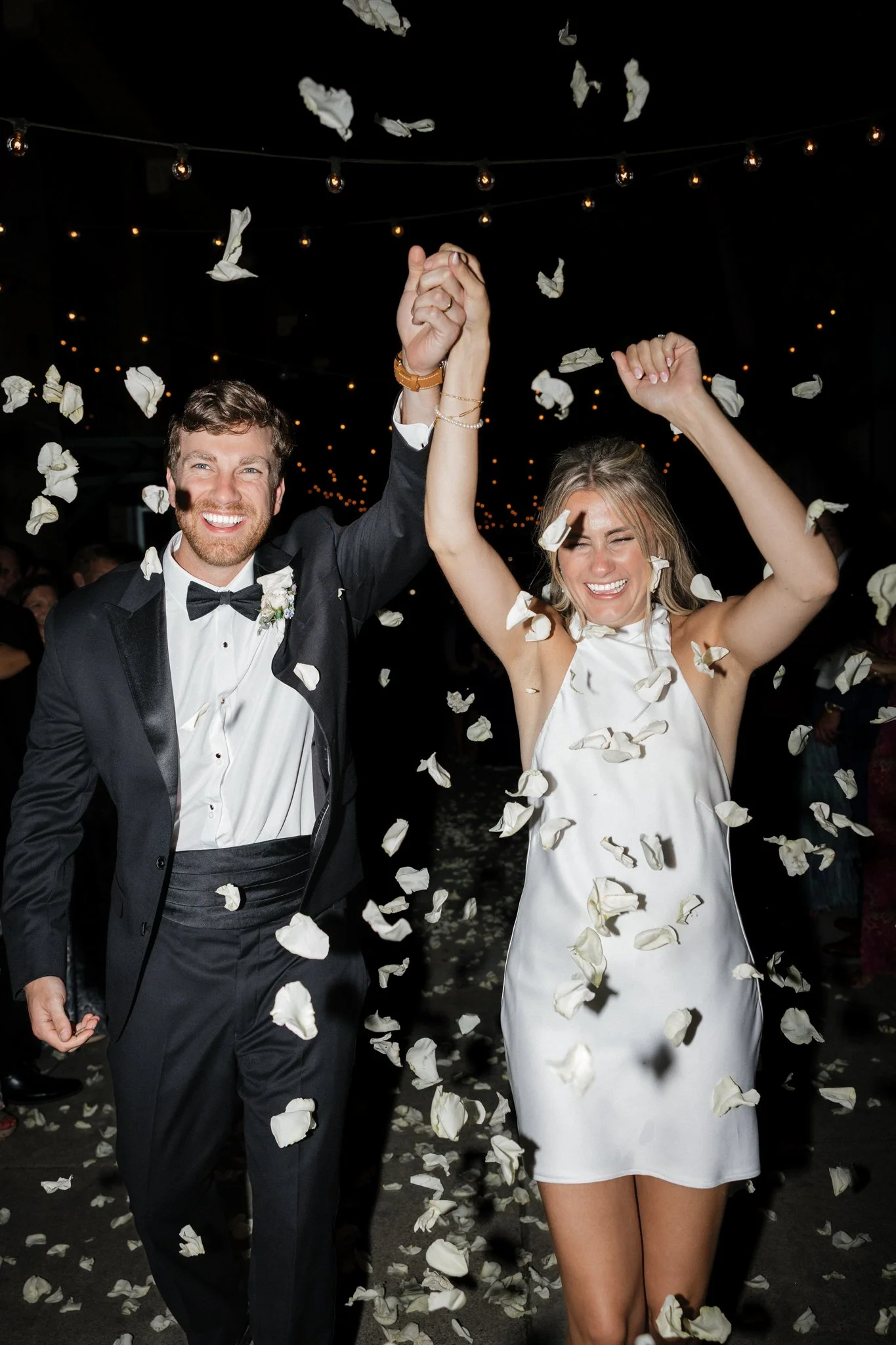 A newlywed couple joyfully celebrating their wedding reception with falling flower petals around them. The groom is wearing a tuxedo and the bride is in a white dress.