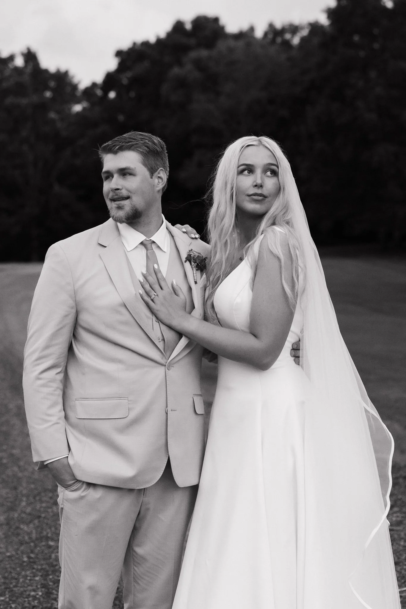 Black and white photograph of a bride and groom standing outdoors on a grassy area, with trees in the background, during their wedding day.