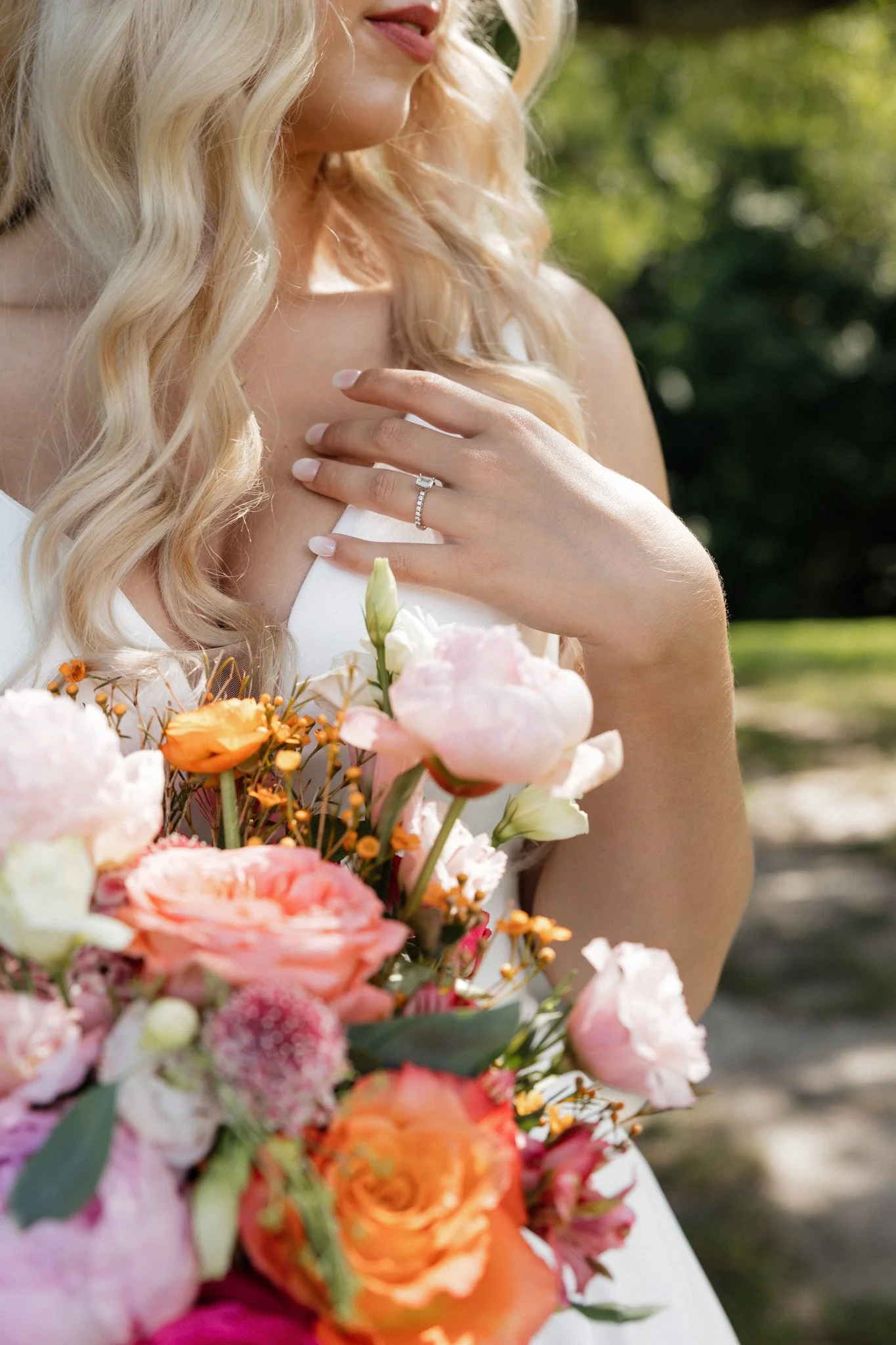 A woman with long blonde hair holding a bouquet of pink, orange, and white flowers wearing a white dress and a diamond ring on her finger.