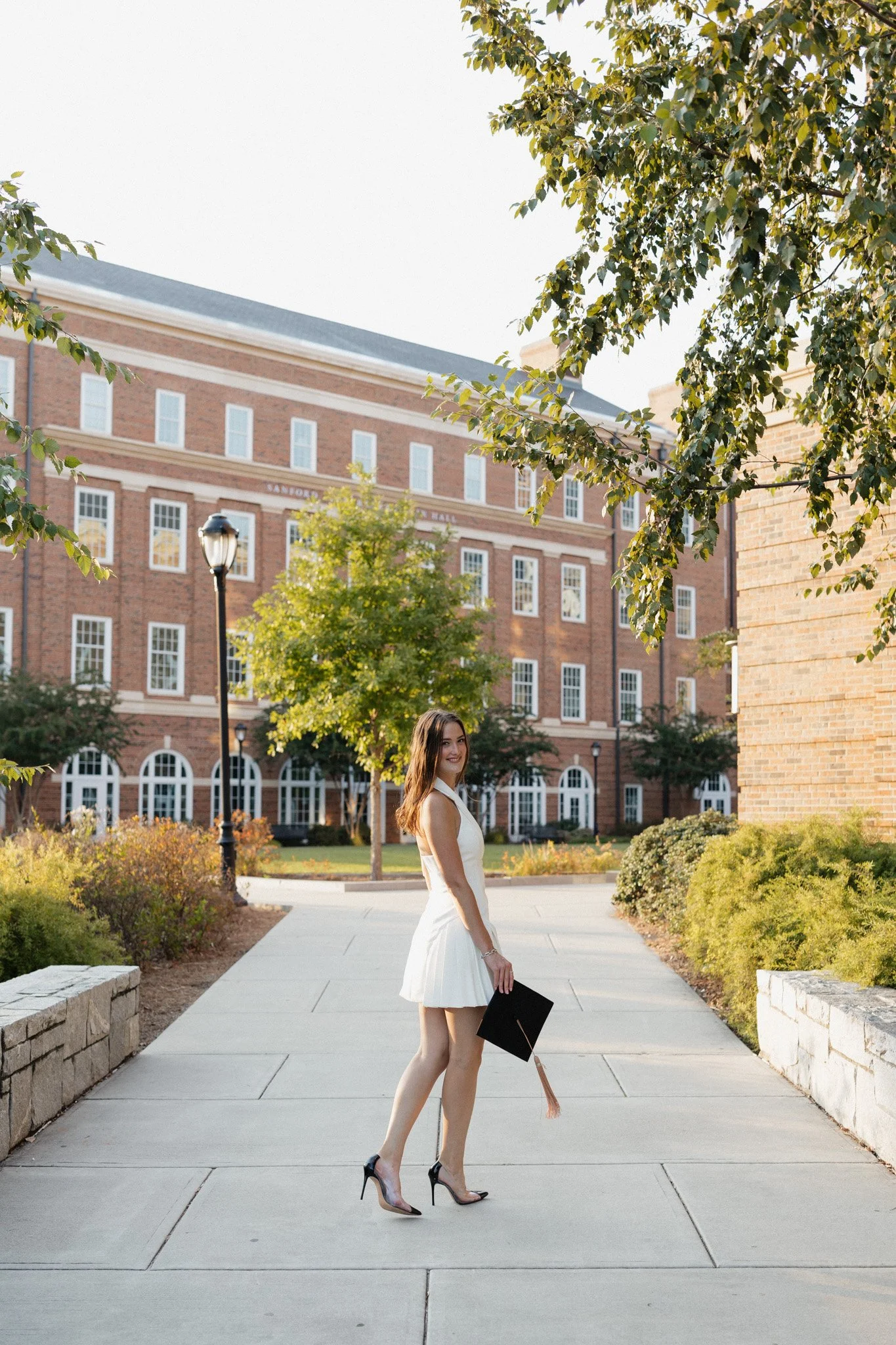 A young woman in a white dress and high heels holding a graduation cap stands on a school campus sidewalk, with a brick university building in the background and trees lining the path.